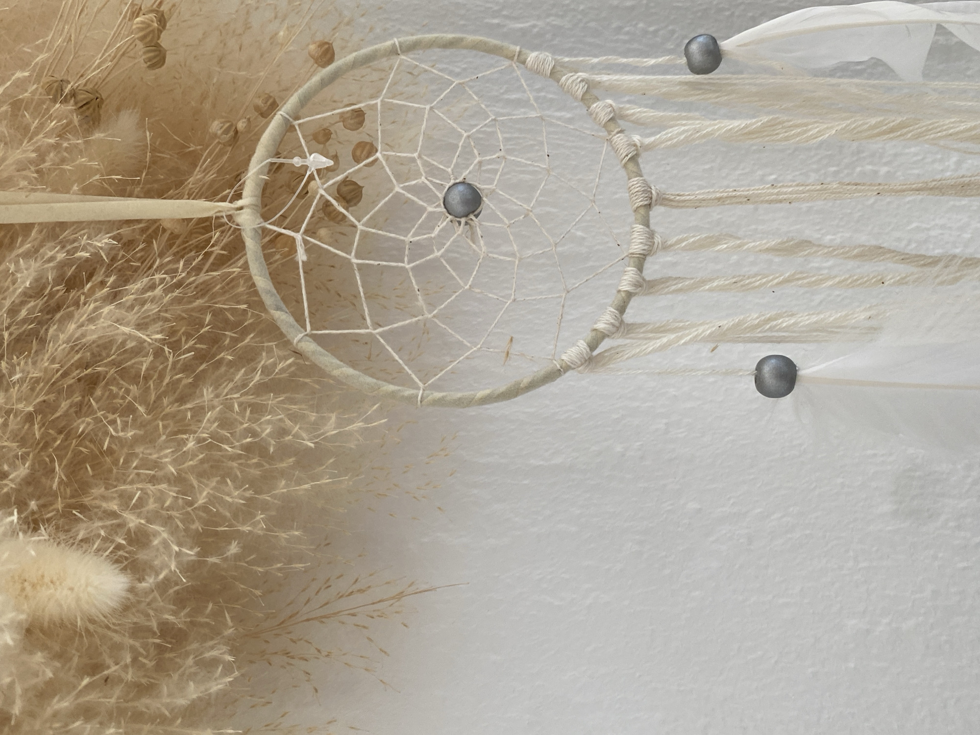 A dreamcatcher made of natural materials hangs against a white, textured wall. The dreamcatcher has a circular frame wrapped in light-colored yarn, with a web of white string inside. A single dark gray bead is suspended in the center of the web. Several strands of yarn hang from the bottom of the frame, each adorned with a dark gray bead and a white feather. The background is filled with dried grasses and seed pods, creating a warm, earthy aesthetic. This object is displayed indoors, likely as a decorative item. No specific location in Bad Sachsa, Germany is identifiable from the image.