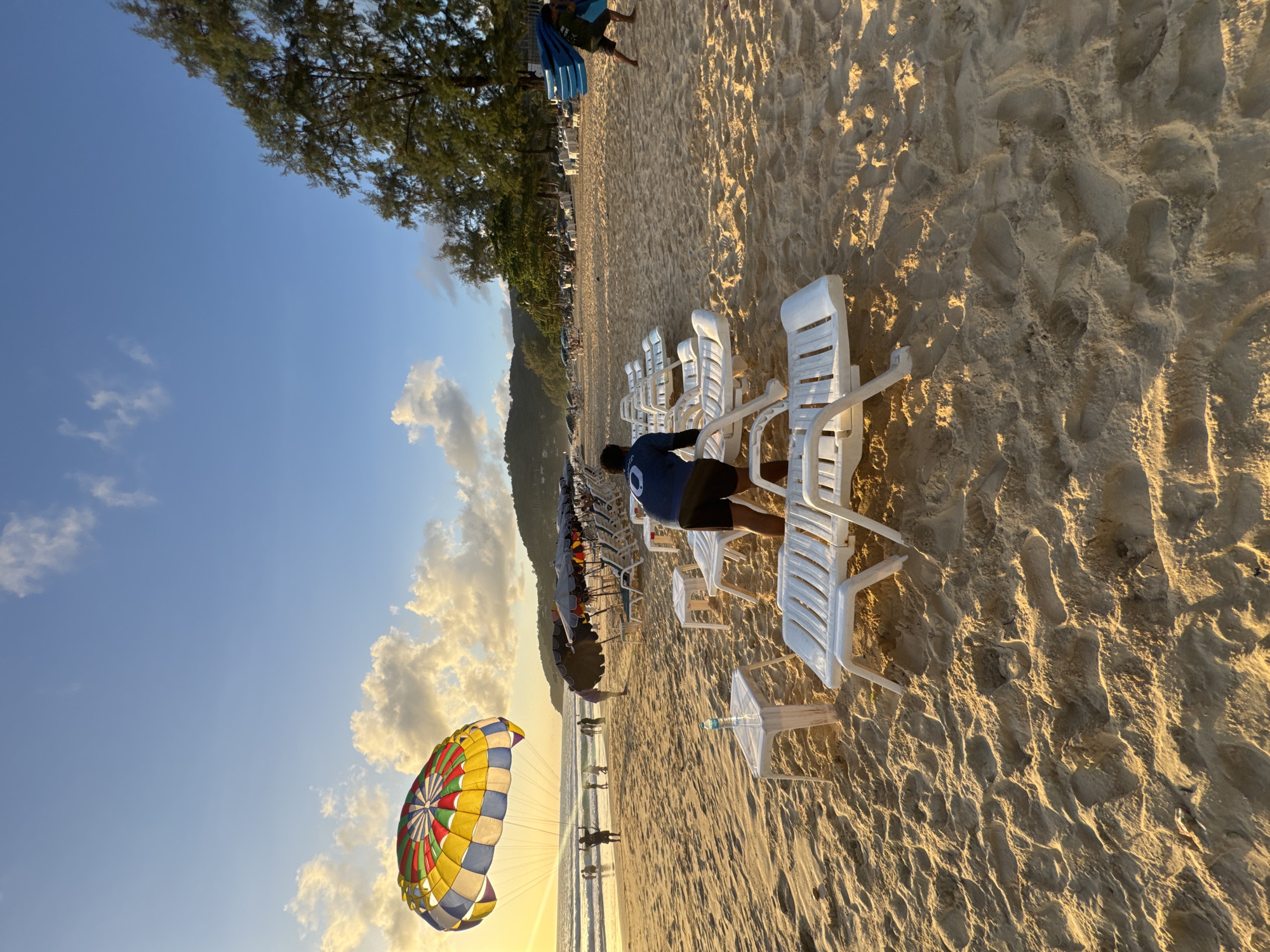 A wide-angle, low-angle shot captures the sandy expanse of Karon Beach in Thailand at sunset. The sun, low on the horizon and to the left of the frame, casts a warm glow across the sand, highlighting the footprints and textures. A colorful parasail, brightly striped in red, yellow, blue, and green, floats in the sky to the left, tethered by lines to a person standing on the beach. In the foreground, several white plastic lounge chairs are arranged in rows, with a small white table and a bottle of water on one.
Further back, a person in a dark blue shirt and black shorts is tending to the lounge chairs. Beyond them, a line of closed beach umbrellas shades more empty chairs. In the distance, gentle waves roll onto the shore where several figures can be seen walking or standing. The background features a line of trees and hills under a sky filled with scattered white clouds and the soft golden light of sunset. On the far right, another person is carrying several folded blue mats. The overall scene depicts a serene beach setting as the day winds down, with activities like parasailing and lounging evident.