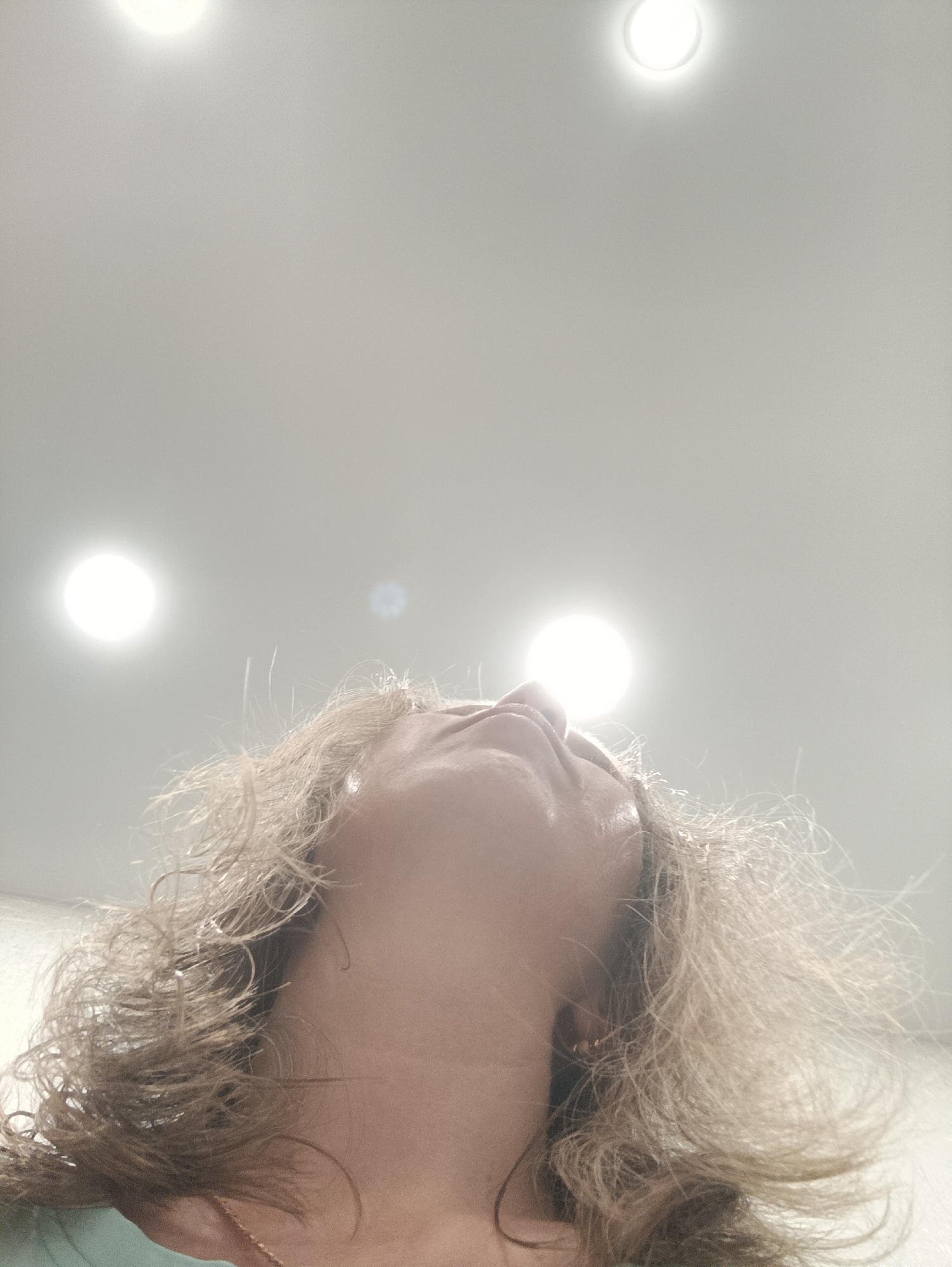 The image is taken from a low angle, looking upwards at a person's face and the ceiling. The person has curly blonde hair and is wearing a light-colored top. The ceiling is white and has three recessed lights, each with a circular shape. The location cannot be determined from the image, and there is no indication it is in Chaikovsky, Russia.