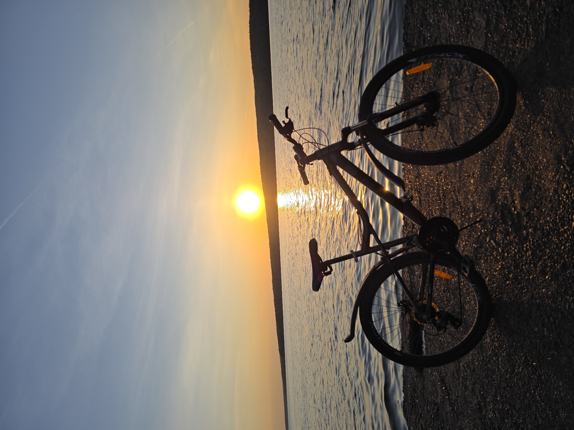 An outdoor scene captures a bicycle positioned on the edge of a body of water, likely a lake or large river. The sun is setting on the horizon, creating a vibrant golden hue across the water and sky. A strip of trees can be seen in the distance, separating the water from the skyline. The bicycle is angled in the foreground, with the water and sunset in the background. The bike rests on the shoreline, which consists of pebbles. The location is plausibly near the city of Chaikovsky, Russia, based on geographic features. There are no people present in the image.