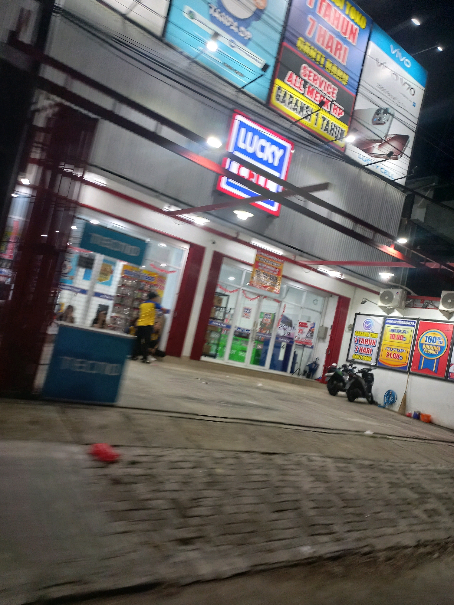 The image depicts a street-level view of a "Lucky" branded store, likely a mobile phone or electronics retailer, in Depok, Indonesia. The store's facade features prominent signage, including a blue and red "LUCKY" logo and advertisements for mobile phone services and brands like Vivo. The store appears to be open, with lights on inside and customers visible through the windows, including individuals at a service counter. Outside the store, a few motorcycles are parked on the pavement, suggesting a common mode of transportation in the area. The environment is dimly lit, indicating it is nighttime or late evening, with artificial lighting from the store and streetlights illuminating the scene. The overall atmosphere suggests a busy commercial area in a Southeast Asian city.