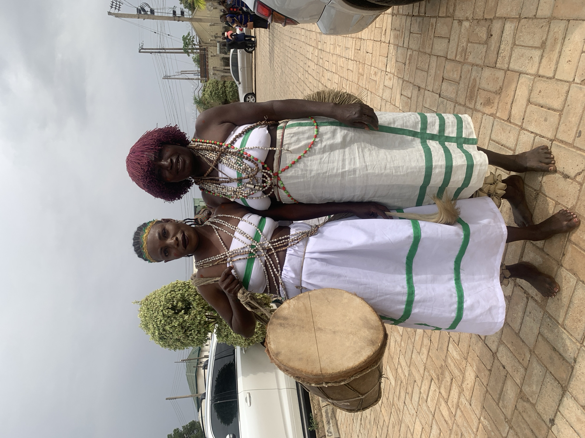 Two dark-skinned women, barefoot and adorned with white fabric dresses with green stripes, and necklaces of white and colored beads, stand on a brick pavement in Jos, Nigeria. The woman in the foreground holds a drum. The background reveals power lines, vehicles, and buildings, suggesting an urban area. The sky is overcast.