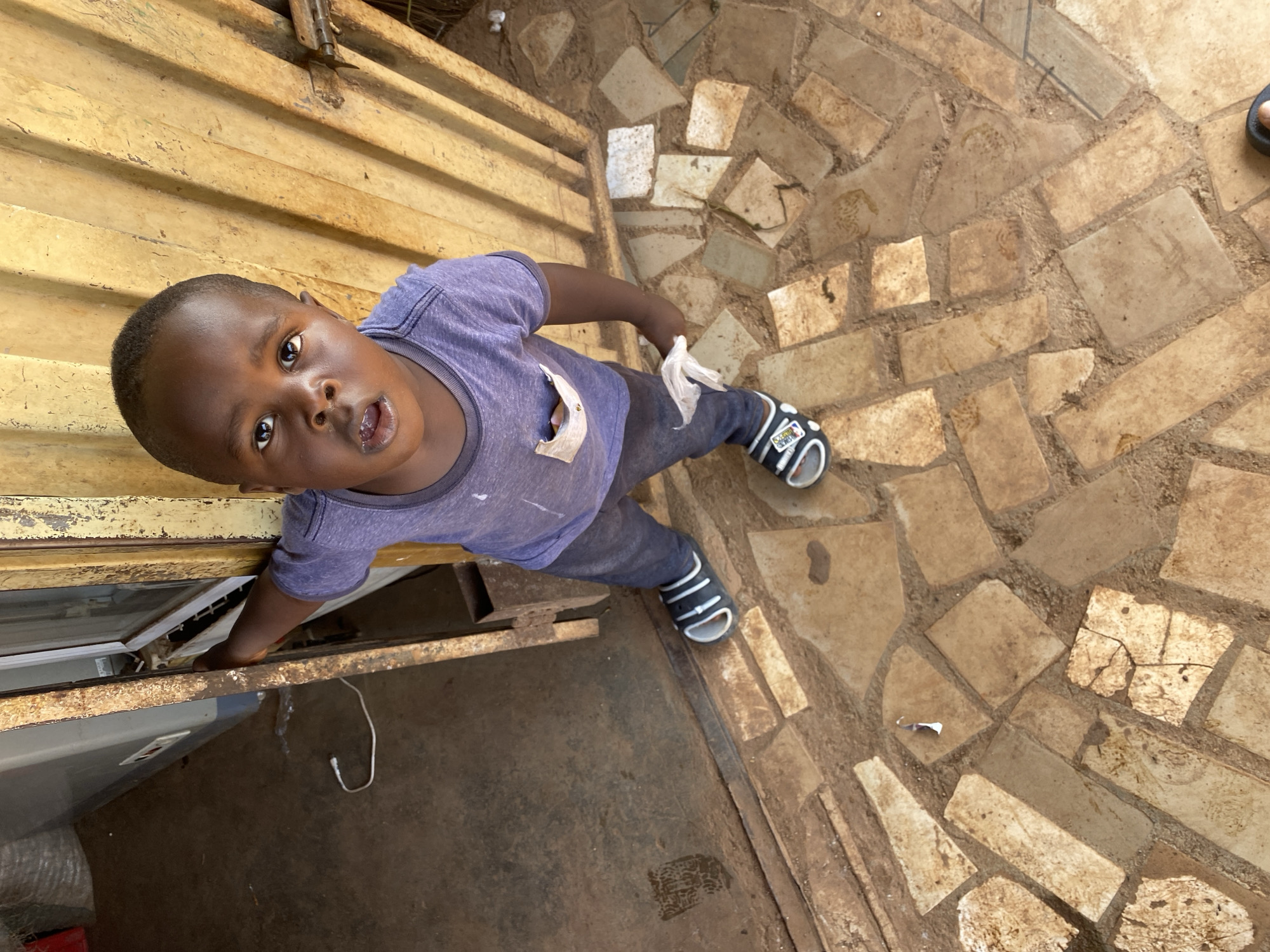 An overhead, high-angle shot captures a young Black boy in an outdoor or semi-sheltered setting in Bukuru, Nigeria. He looks directly up at the camera with a curious, open-mouthed expression. The boy is wearing a purple t-shirt, dark trousers, and black and white sandals; the strap of his right sandal clearly displays the text "MADE IN".
His right arm is extended downwards, reaching towards a white cooler or appliance situated next to a dark, unpaved area. Above the cooler, a weathered, yellow-brown corrugated metal structure, possibly a wall or counter, shows signs of rust and features a visible metal latch in the upper background. The ground around the boy is paved with irregular, light brown flagstones set into darker earth or mortar, forming a rustic mosaic pattern. A small piece of white paper or plastic debris lies on the paved ground, and a white cable is visible on the darker, unpaved section near the cooler. A partial view of another person's foot is visible in the far right of the frame. The scene is brightly lit, indicating it is daytime.