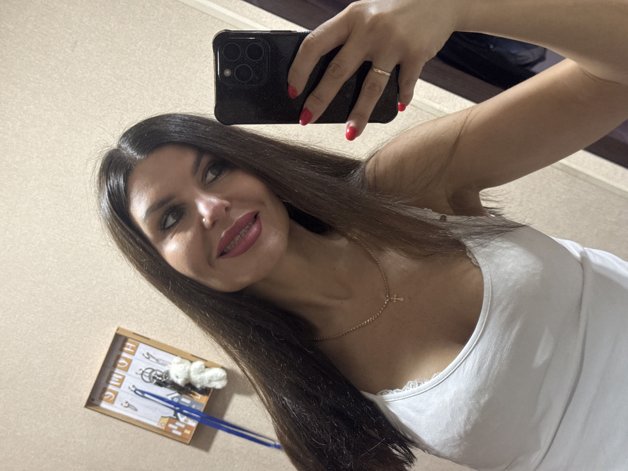 A woman is taking a selfie in a room with beige walls. She has long, dark hair and is wearing a white tank top. She is also wearing a necklace with a cross pendant and has red nail polish on her fingernails. In the background, there is a framed key holder with the word "HOME" and some keys and a small white teddy bear hanging from it.