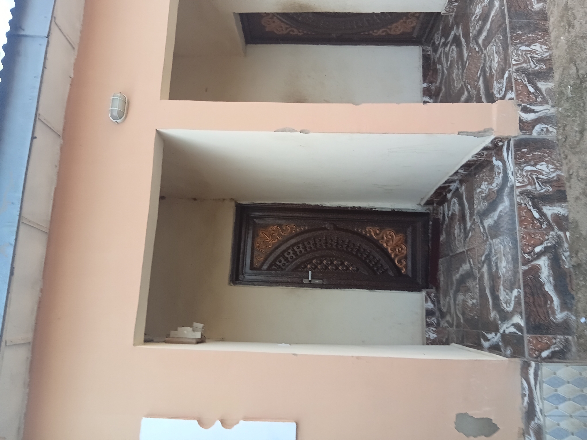 The image displays a detail of an outdoor space, possibly a building entrance or walkway in Jalingo, Nigeria. The primary subjects are architectural elements and decorative features. A peach-colored wall dominates the left side, with a white, fluted outdoor light fixture attached to it. To the right of the wall, there is an opening revealing an interior space with a dark, intricately carved wooden door. The door features ornamental patterns with gold accents and a small metal latch. Above the door, a section of the ceiling or upper wall is visible, also decorated with a similar dark wood and gold pattern.
The flooring adjacent to the peach wall is tiled with a marbled pattern of brown, white, and dark red. Further to the right, another tiled area shows a geometric pattern of blue and grey diamonds. The scene appears to be well-lit, suggesting daylight, though no specific time of day or weather conditions are evident. The overall impression is of a functional outdoor area with decorative embellishments. There is no text visible in the image.