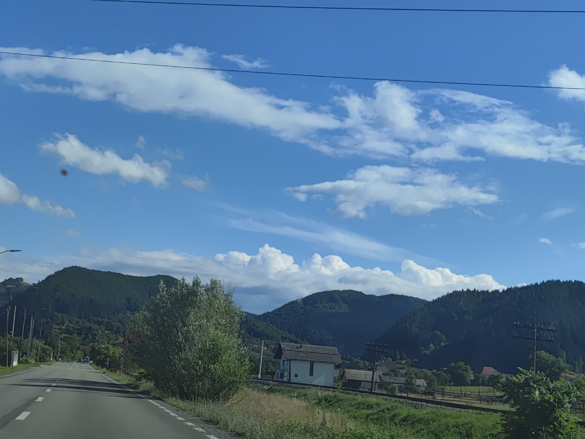 A view from a car on a paved road in Leordina, Romania, shows a rural landscape with mountains, trees, and a few buildings. The road curves to the left in the foreground, with white dashed lines marking the lanes. On the right side of the road, there is a grassy area with some trees and bushes. Further back, a white house with a dark roof is visible, along with other structures and a railway track. The background is dominated by green, tree-covered mountains under a bright blue sky with scattered white clouds. Overhead power lines are visible in the upper portion of the image. The lighting suggests it is daytime, likely midday.