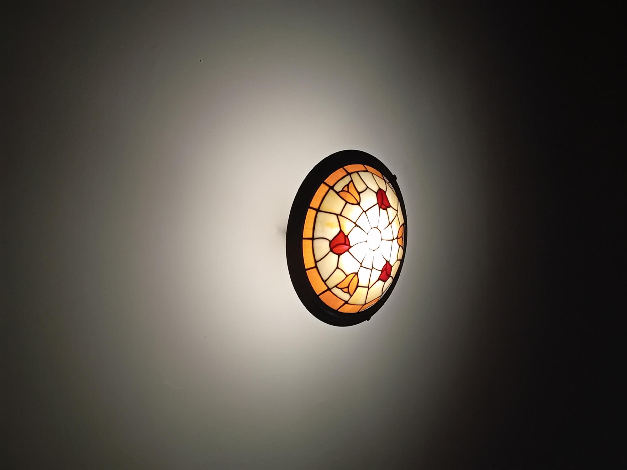 An image displays a round, illuminated light fixture mounted on a wall or ceiling, centered slightly to the right. The fixture is a stained-glass lamp, possibly in a Tiffany-style, featuring a dark, circular frame. The glass panels within the lamp form a mosaic pattern, with predominant colors of cream or off-white, warm orange, and a deep red. Some of the red and orange pieces are shaped like stylized leaves or petals, arranged symmetrically around the central light source.
The lamp emits a warm, bright glow that strongly illuminates the immediate surrounding surface, creating an oval-shaped patch of light. Beyond this illuminated area, the background transitions smoothly into a dark, gradient gray and black, indicating that the lamp is the primary source of light in a dim or dark interior space. This suggests it is either night or a room with low ambient lighting.
No people, specific activities, or discernible emotions are present in the image. There are a few very small, faint specks visible in the lighter areas of the background, likely dust particles or minor lens imperfections. No text is visible anywhere in the image. The focus is entirely on the decorative light fixture and its luminous effect on the surrounding environment.