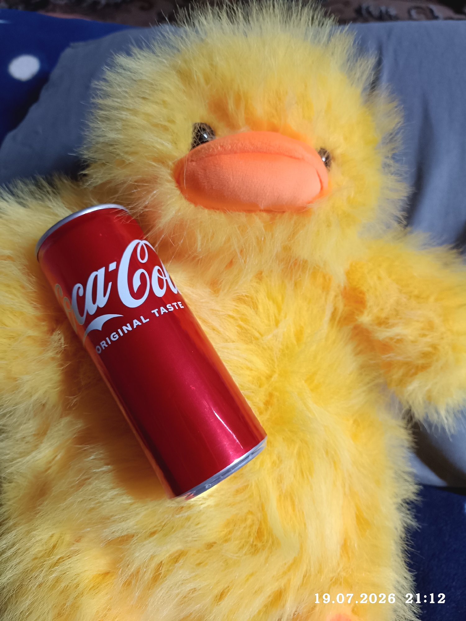 A close-up, slightly elevated, top-down view shows a metallic red Coca-Cola can resting on a bright yellow, fluffy stuffed duck. The duck's orange beak and dark eyes are partially visible in the upper center of the frame. The can, angled diagonally from the bottom left to the upper right, displays the white Coca-Cola logo and the text "ORIGINAL TASTE". The background consists of a blurred dark blue fabric with white circular patterns, and a grey fabric. A timestamp in the bottom right corner reads "19.07.2026 21:12". The image does not provide any information about the location in Radisne, Ukraine.