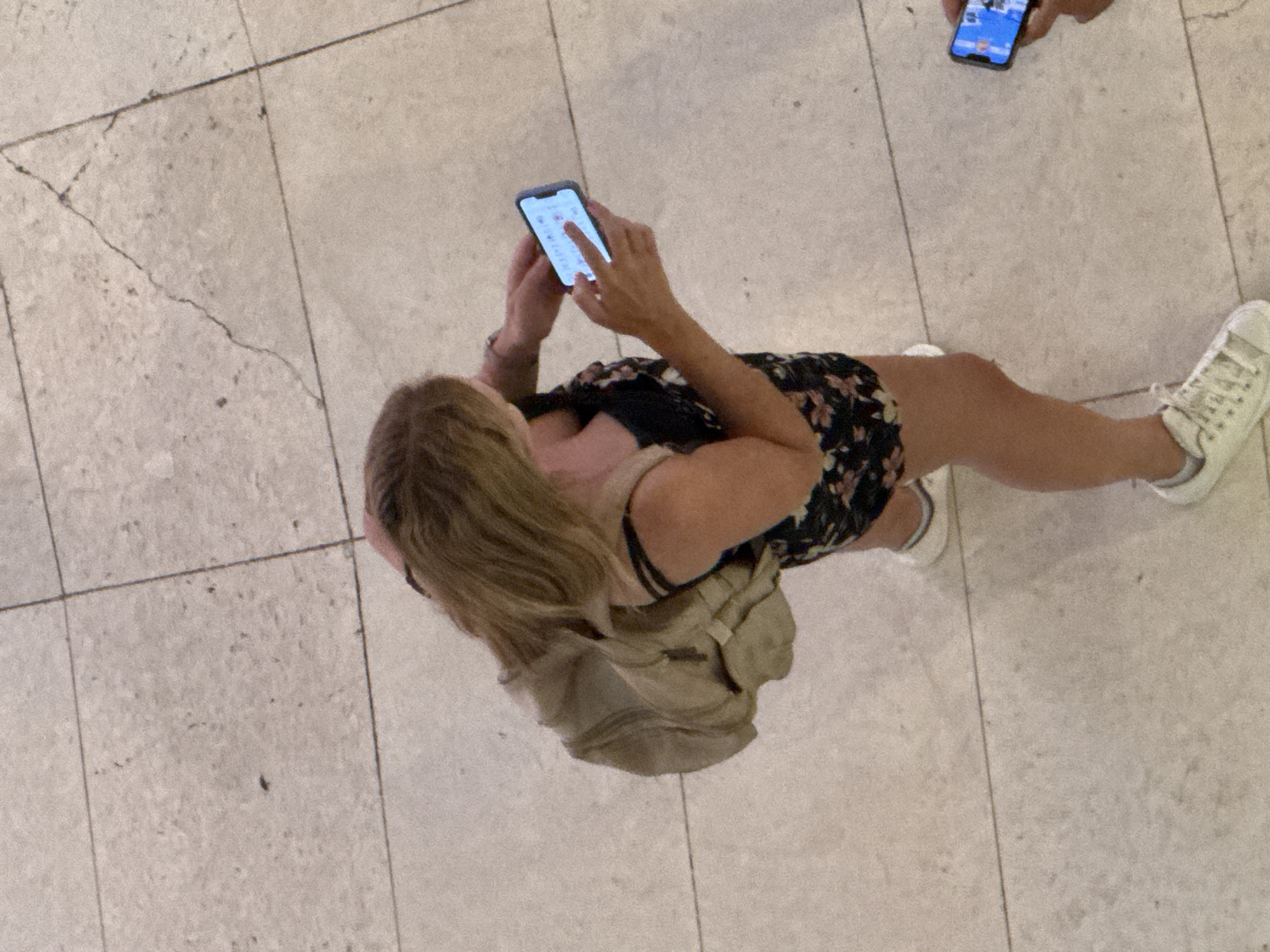 This image features two individuals interacting with their smartphones on a tiled floor, likely indoors or in a sheltered outdoor area given the lighting. The primary subject is a woman viewed from above, standing and looking at her phone. She has blonde hair and is wearing a black floral-print dress with thin straps and a tan backpack slung over her shoulder. Her right hand is raised, and her index finger appears to be touching the screen of her phone, suggesting she is actively using it, possibly texting, browsing, or playing a game. The phone is silver and held in a vertical orientation.
To the right and slightly above the woman, another person's hand and smartphone are visible. This phone displays a blue screen with some icons or text, also being held in a vertical position. The perspective suggests this second person might be standing or walking.
The setting is a floor made of large, light-colored square tiles, with thin dark grout lines dividing them. Some tiles show minor cracks and imperfections. The lighting is bright and somewhat diffused, creating subtle shadows. There are no clear indications of the time of day or weather. The overall impression is one of everyday activity, with people engrossed in their personal devices. The location context of Barcelona, Spain, is provided but not visually evident in the cropped image. No discernible text is readable on the phones or in the scene.