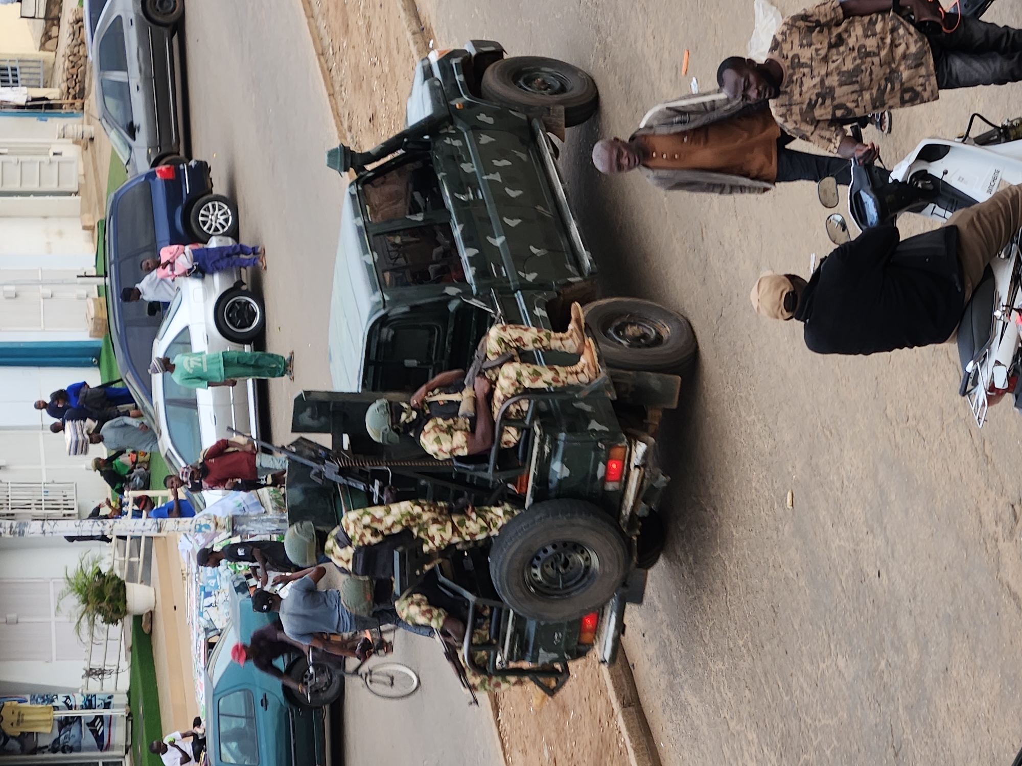 In an outdoor urban setting, a military vehicle, a dark green four-door pickup truck with a camouflage pattern and what appears to be a mounted machine gun, is parked on an asphalt road. Three individuals in camouflage military uniforms are seated in the back of the truck, one holding a rifle. Two of them are wearing helmets. A fourth individual in a camouflage uniform is standing near the truck, holding a rifle. In the foreground, a man in a brown shirt and dark pants walks past the truck, and two other men are visible near a white scooter; one is wearing a tan baseball cap and black jacket, while the other wears a camouflage t-shirt and dark pants. Several cars are parked in the background, along with a building and trees.