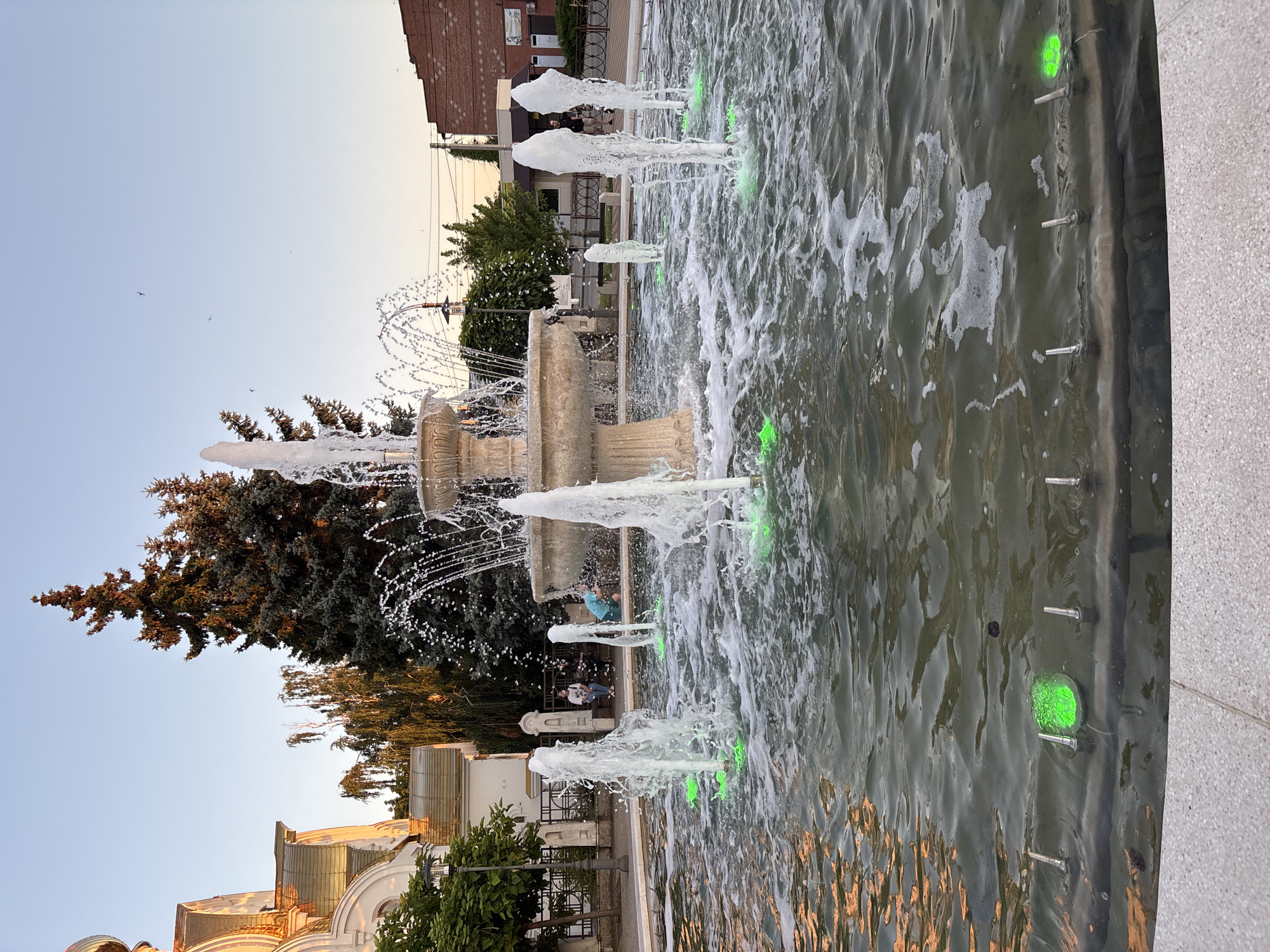 The image shows a multi-tiered fountain with several water jets spraying upwards, creating a frothy effect in the water. The fountain is illuminated with green lights from below the water's surface. In the background, a building with a golden dome and white walls is visible, suggesting a church or religious structure. Evergreen trees and other foliage are present, along with some urban infrastructure like buildings and a fence. A few people are seated on a bench in the middle ground, and one person is standing near the fountain in the background. The sky is clear and light blue, indicating daytime. The scene appears to be a public square or park area within the city of Tbilisskaya, Russia.