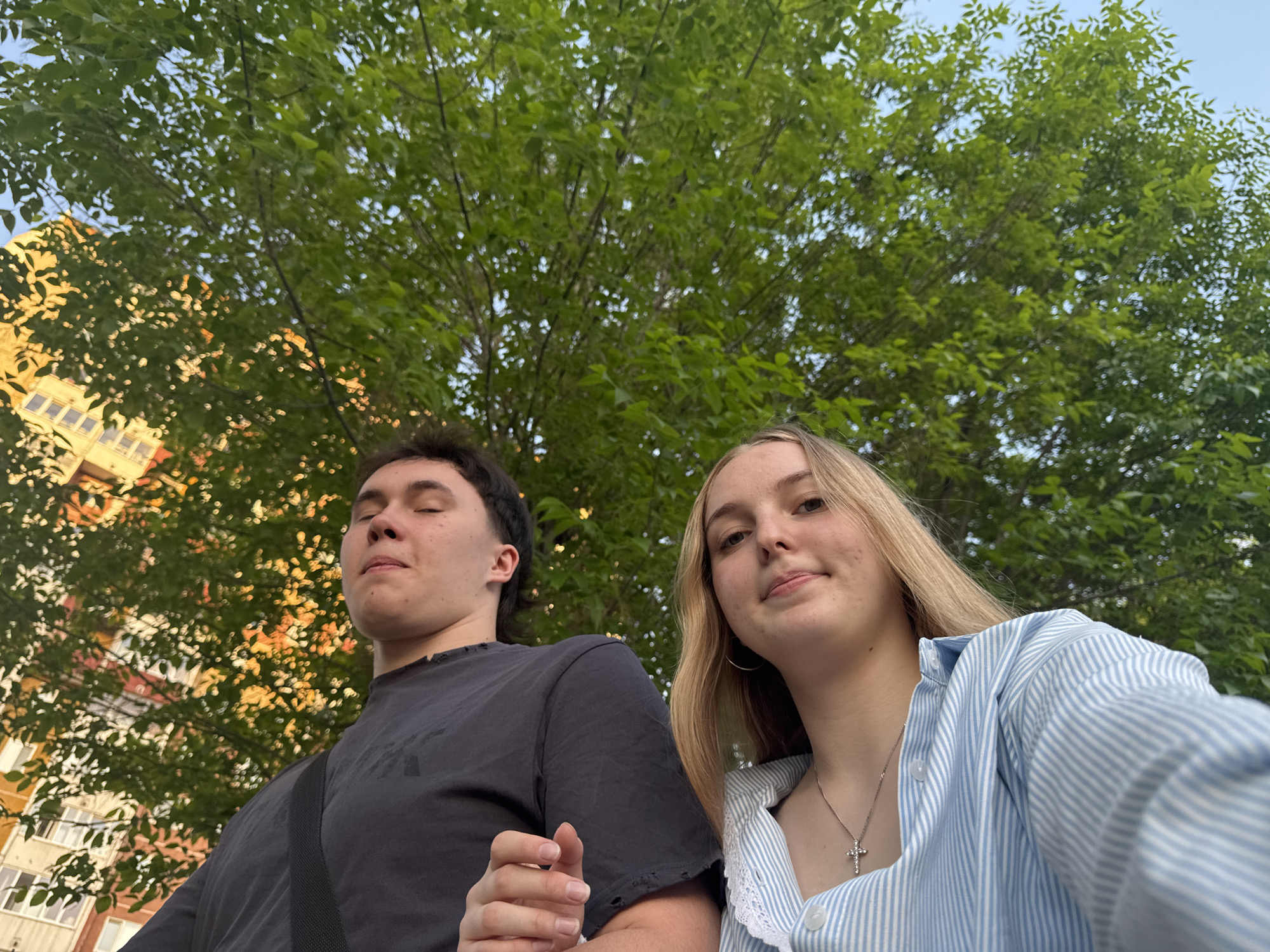 Two young people, a male and a female, are posing for a selfie in front of a large tree with green foliage. The male is on the left wearing a dark-colored t-shirt, while the female is on the right wearing a light blue and white striped shirt with a silver necklace. Behind them, there's a building with a yellow facade. The photo was taken in Perm, Russia.
