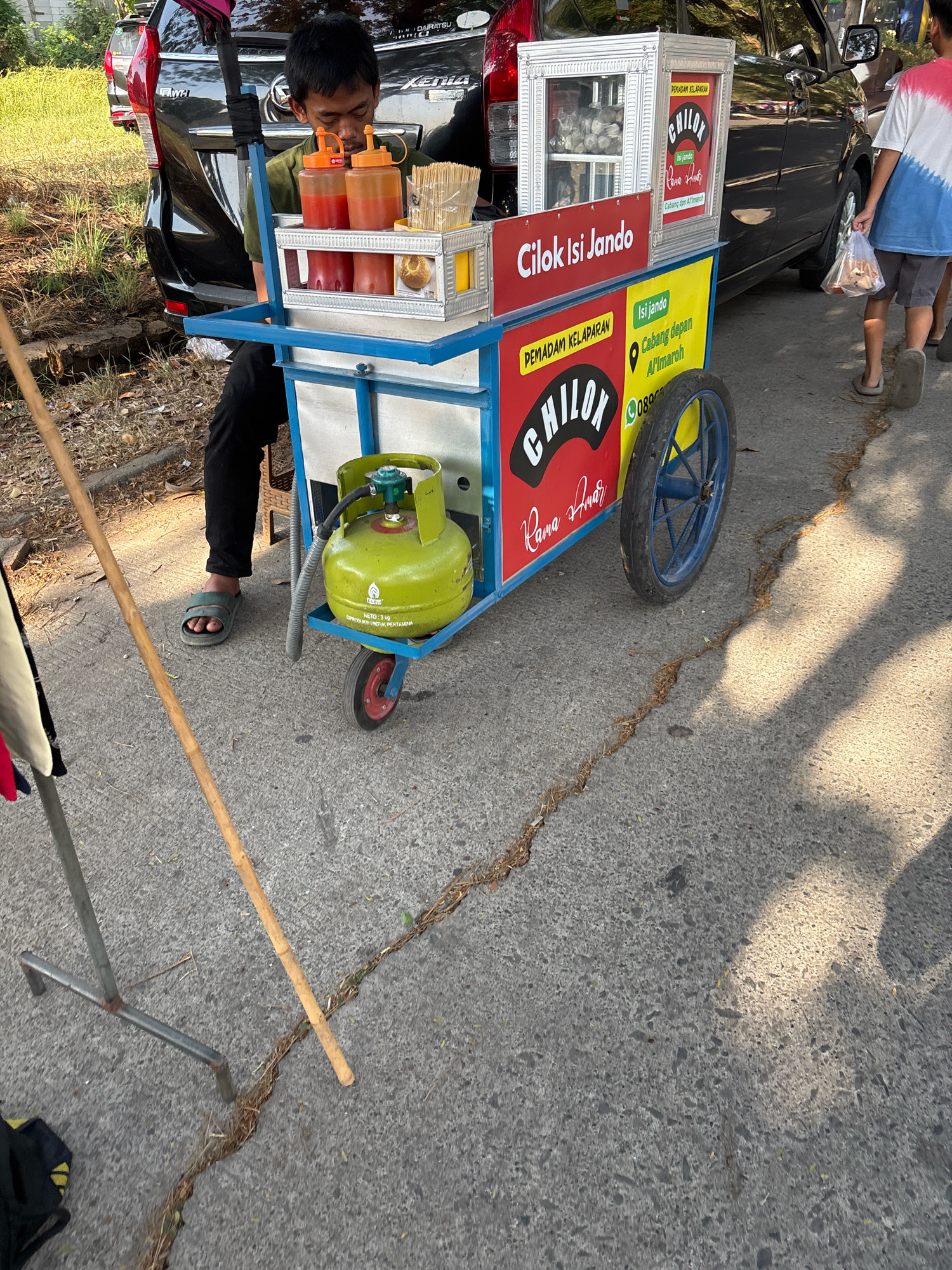 In Telagamurni, Indonesia, a young man is seated behind a mobile food cart selling "Chilok Isi Jando." The cart is vibrant, with a yellow and red body and blue trim, and it displays advertising banners for the food product. A green gas cylinder is attached to the cart, indicating its operational setup. Two bottles of sauce, presumably chili and tomato, are positioned on top of the cart, along with a container of wooden skewers. In the background, a black car, possibly a Daihatsu Xenia, is parked. A child wearing a tie-dye t-shirt and shorts walks away from the cart, carrying a plastic bag, suggesting a customer interaction. The scene unfolds on a paved road with a visible crack running through it, lined with dry vegetation. Sunlight casts shadows across the ground, indicating a sunny day. A metal stand and a wooden pole are also present in the foreground.