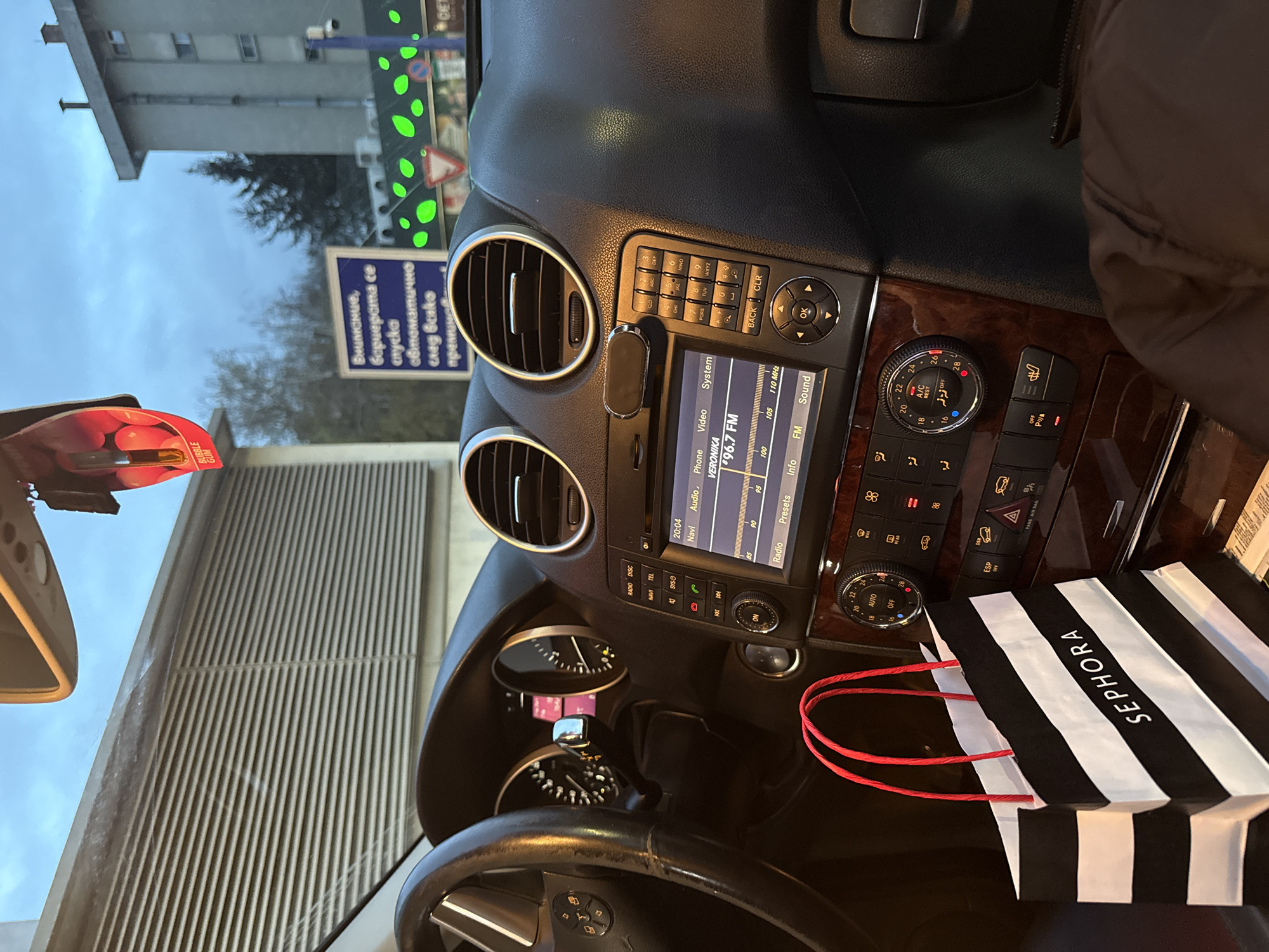 The image captures the interior of a car, specifically the dashboard and part of the steering wheel. The car's infotainment system displays "VERONIKA" and "96.7 FM," indicating a radio station. To the right of the infotainment system is a Sephora shopping bag with red handles. Part of a red bubble gum pack is visible on the left. Through the windshield, we see a building and a sign with the text "Внимание, бариерата се спуска автоматично след всяко преминаване," which translates to "Attention, the barrier lowers automatically after each passage". This sign is written in Bulgarian, confirming the location is in Bulgaria. Based on the architectural style of the building, the environment and the sign being in Bulgarian, the location is likely in Sofia, Bulgaria.