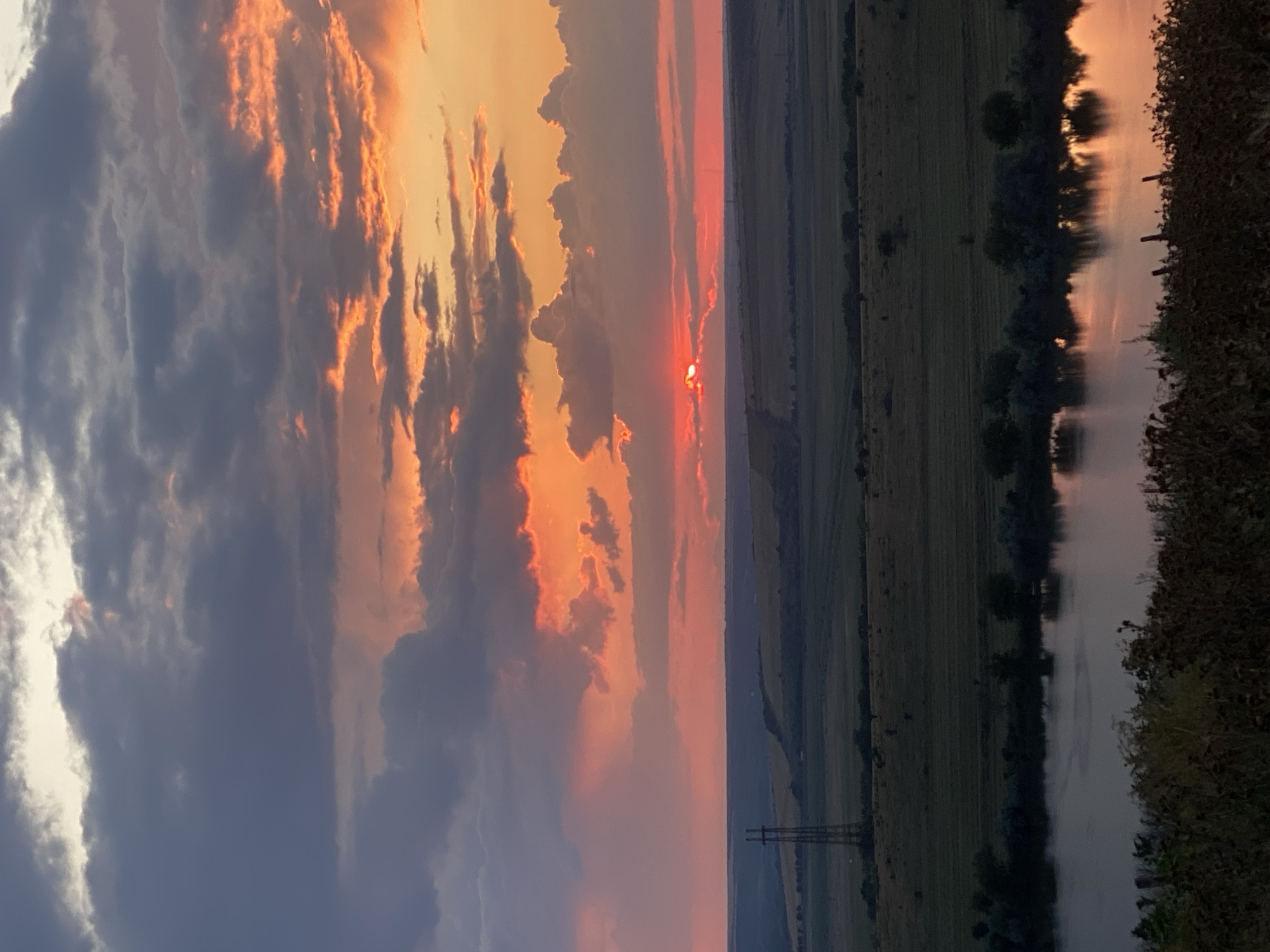 A sunset over a rural landscape in Cairaclia, Moldova. The sky is filled with dramatic clouds tinged with vibrant orange and red hues. The sun, a small bright orb, is partially obscured by clouds near the horizon. Below the sky is a vast expanse of rolling fields, some appearing cultivated. A tall power line tower stands prominently in the middle ground. In the foreground, a body of water reflects the colors of the sunset, with the shoreline lined by trees and vegetation. The overall scene depicts a serene natural environment at dusk.