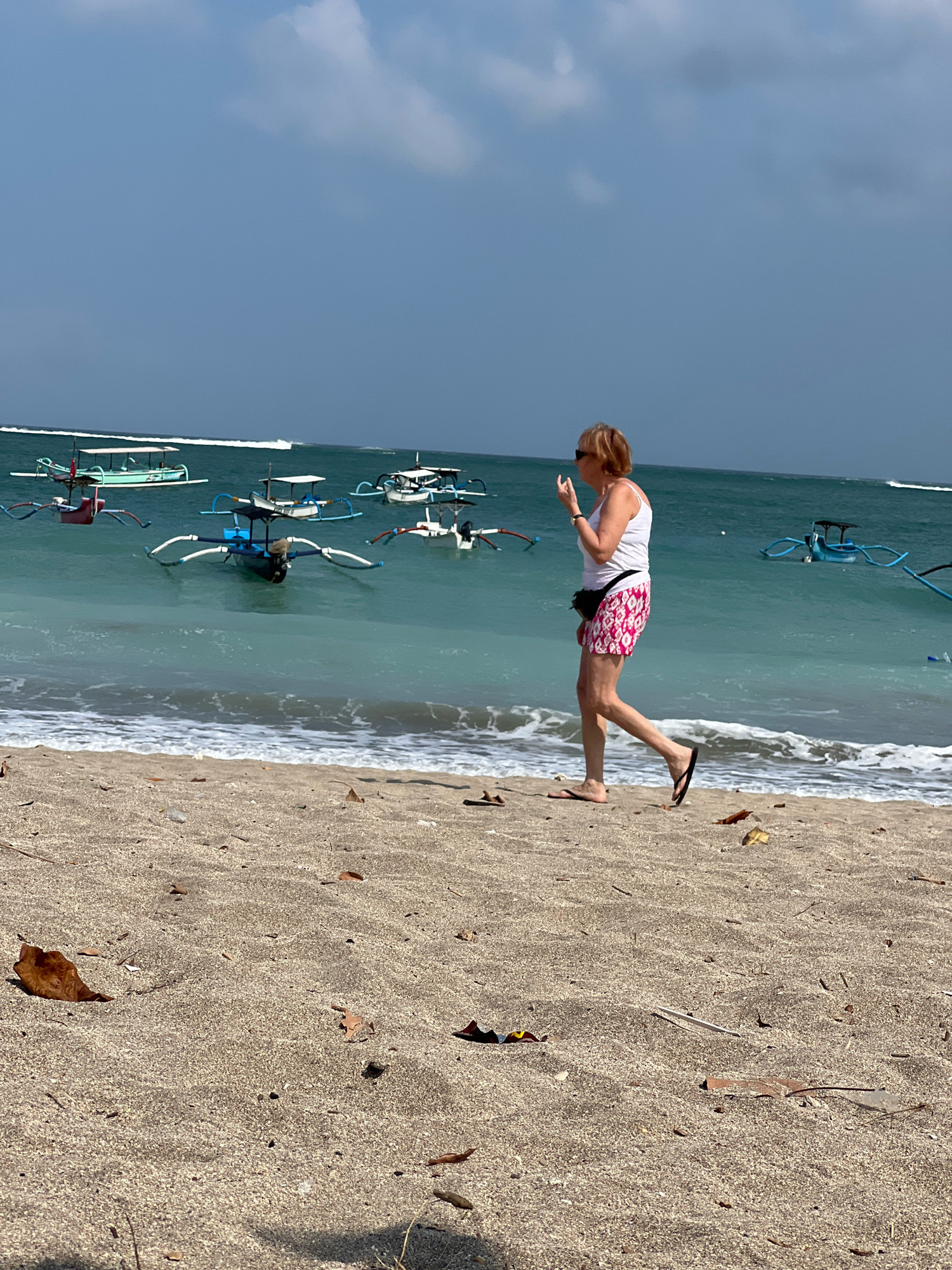 A woman walks on a sandy beach in Kuta, Indonesia. She is wearing a white tank top and pink and white patterned shorts, with a black fanny pack around her waist. She is wearing sunglasses and appears to be looking at her phone as she walks. Her left arm is bent, and her hand is near her face, possibly holding the phone. She is wearing flip-flops. The beach is covered in sand with scattered leaves and debris. The ocean is visible in the background with several traditional Indonesian fishing boats, known as jukung, dotting the turquoise water. The sky is partly cloudy. The waves are gentle, creating a white line of foam along the shore.