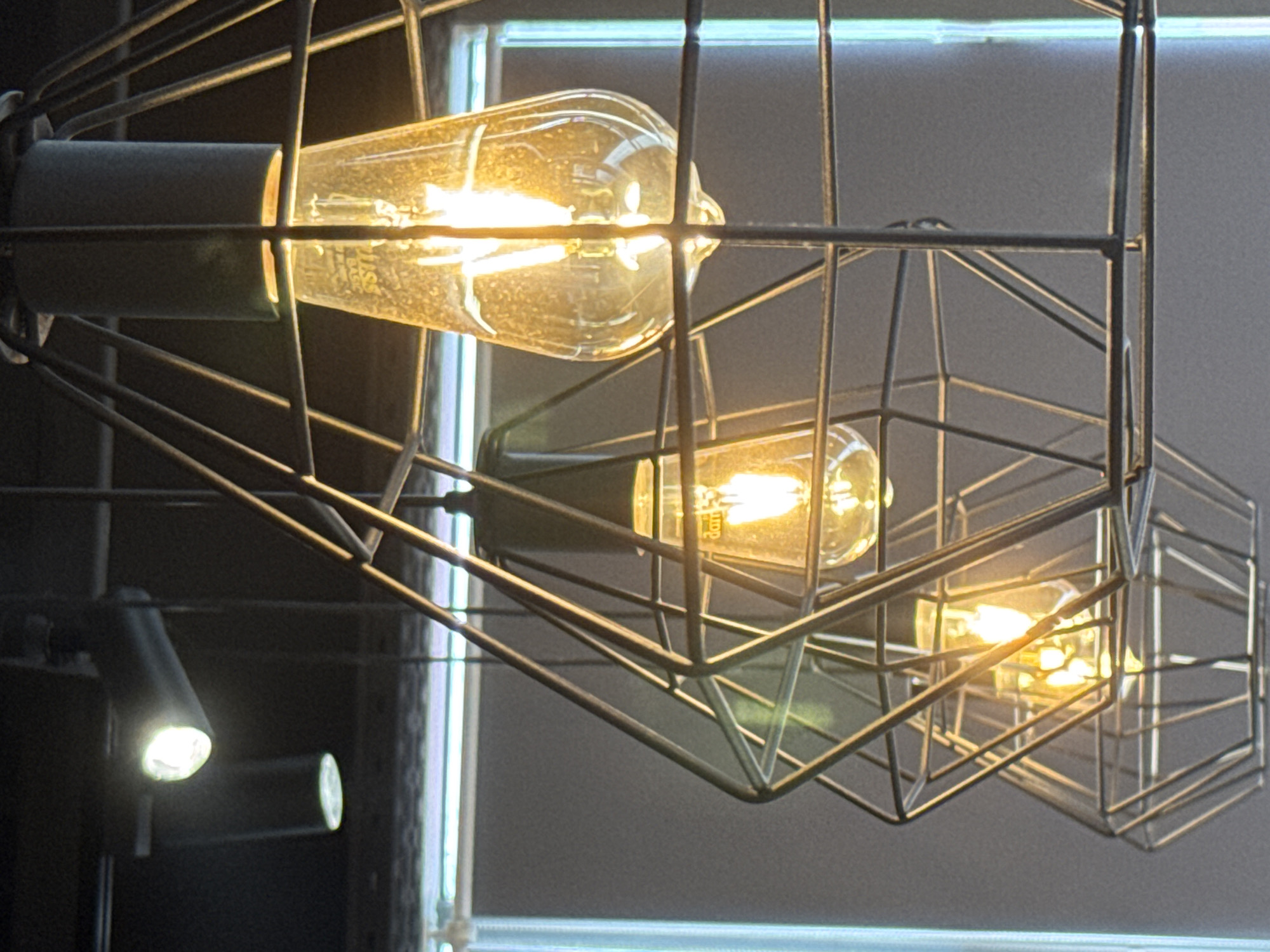 The image shows a close-up of several Edison-style light bulbs housed within geometric wire cages. The bulbs are illuminated, casting a warm, yellow light. The cages are made of dark metal and have a diamond-like structure. In the background, a window with a light blue tint and a dark ceiling with a spotlight are visible. The scene appears to be indoors, possibly a restaurant, cafe, or a modern living space, likely in St. Petersburg, Russia, given the context of the request. There are no people or animals depicted in the image, and no discernible actions or interactions are taking place. The focus is entirely on the decorative lighting fixtures.