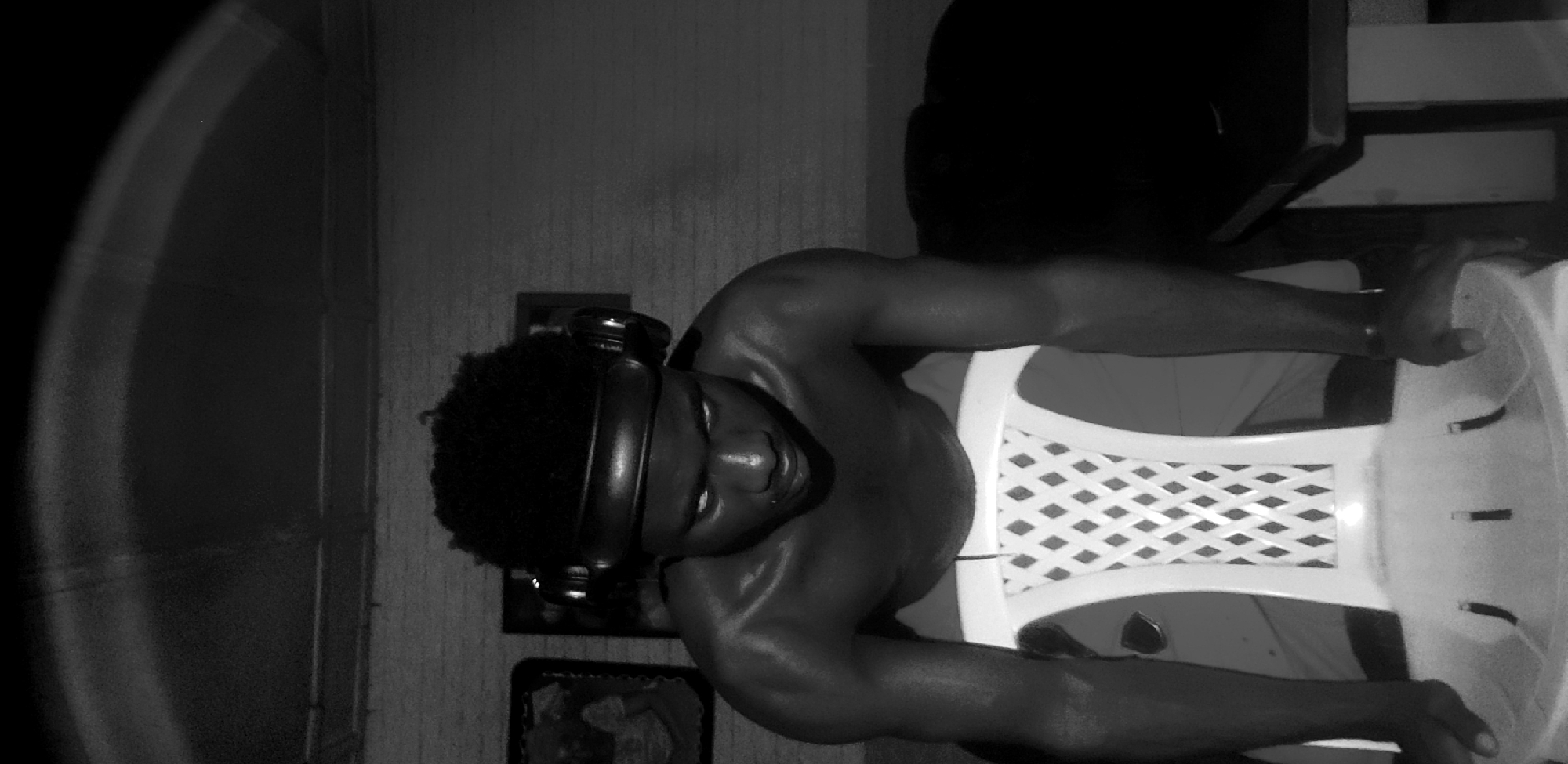 The black and white photograph, likely taken in Narayi, Nigeria, depicts a young Black male from the chest up, leaning forward with his hands on a white plastic chair. He is shirtless, his skin glistening with sweat, suggesting a recent physical exertion or warm environment. The man wears large, over-ear headphones around his neck and on his head. His facial expression is intense, with his eyes wide open and looking downwards, creating a dramatic and focused mood. The background is dimly lit, showing a textured wall, possibly brick, and two framed pictures on the wall. The overall lighting is dramatic, with strong highlights on the man's body and a dark, shadowy environment, contributing to a sense of intensity and perhaps introspection. The image appears to be a portrait focusing on the subject's physical presence and emotional state.