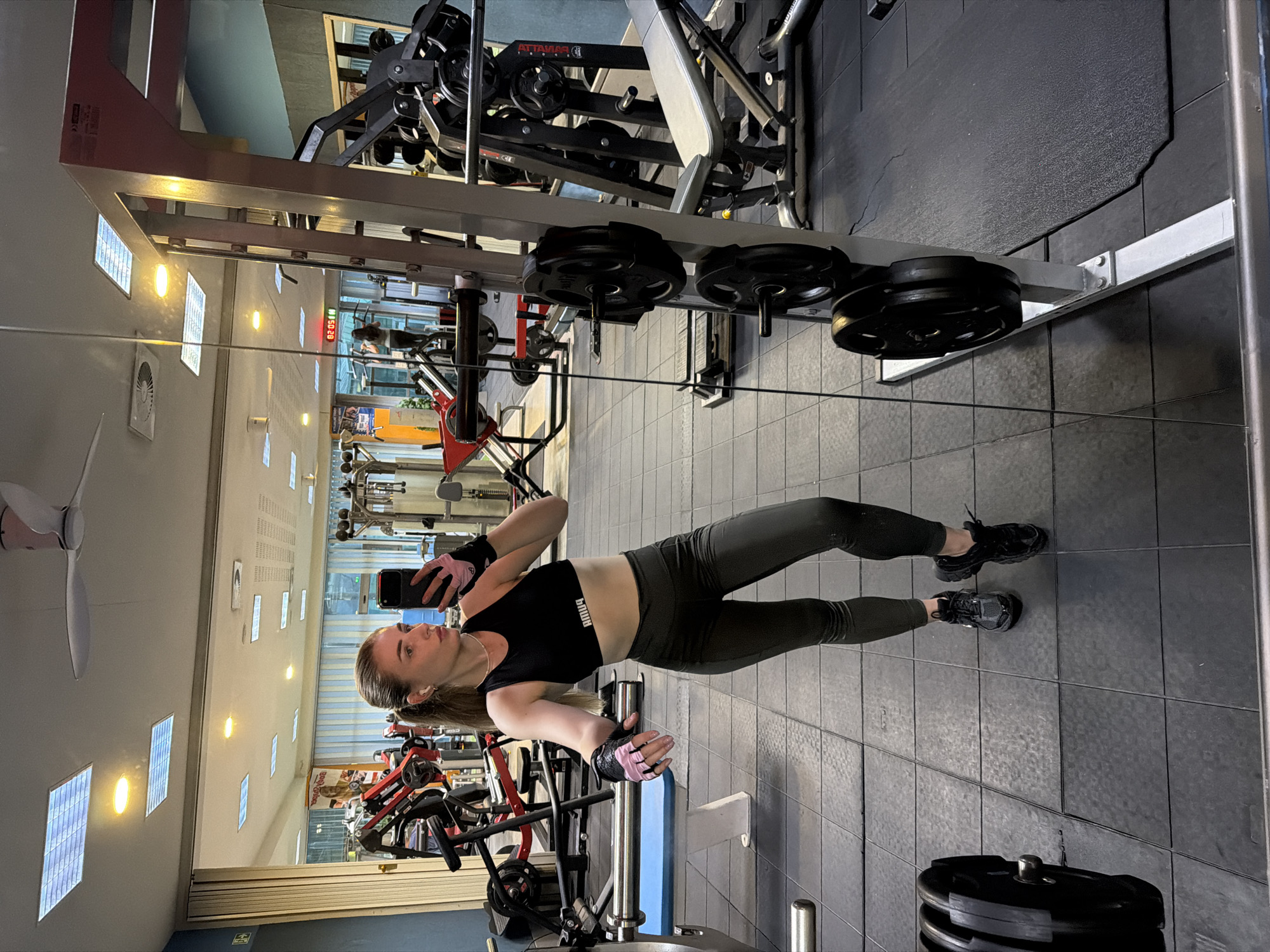 A young Caucasian woman with blonde hair pulled back in a ponytail, wearing a black sports bra, dark grey leggings, and black athletic shoes, takes a selfie in a gym mirror. She is wearing pink and black weightlifting gloves on both hands and small white wireless earbuds. The gym is equipped with various weightlifting machines, including a Smith machine with weight plates attached, a bench press, and other exercise equipment. The floor is tiled with black rubber mats, and the walls are painted in shades of blue and beige. The lighting appears to be fluorescent and overhead. A digital clock in the background shows "02:05".