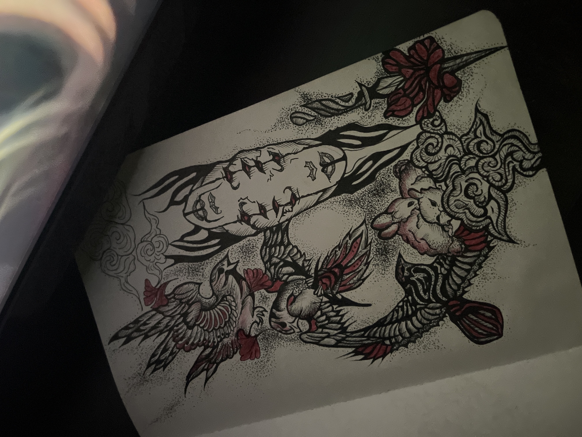 A detailed illustrative drawing is presented on a light-colored sheet of paper, likely from a sketchbook, viewed at an angle. The artwork is executed primarily in black ink with extensive use of stippling for shading and texture, complemented by striking red accents.
The central subject is an elongated, stylized face or mask-like figure, depicted with closed eyes and visible features resembling tears or stitches in red along its cheeks, conveying a sense of sorrow or resilience. Above this figure, a dagger or short sword is prominently displayed, its hilt adorned with a large, intricate red flower or bow.
Several stylized birds, possibly phoenixes or similar mythical creatures, are integrated into the composition, featuring elaborately detailed wings and bodies. These birds also incorporate red coloring on their wings and tail feathers, enhancing the visual flow. They appear to be in dynamic poses, swirling around the central elements.
The design is interspersed with swirling cloud or smoke-like patterns, some showcasing intricate decorative motifs. A small, stylized, almost animalistic head or figure is visible amidst these cloud formations.
The overall composition is dense and dynamic, suggesting a narrative or symbolic theme, possibly involving struggle, emotion, or transformation. No text is visible within the drawing. The photograph is taken in a dimly lit setting, with a dark background surrounding the paper. The context for the photo's capture is Sofia, Bulgaria.