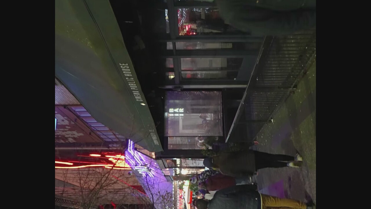 A nighttime, exterior, medium shot captures the entrance to "CINEMA PUBLIC HOUSE" in Vancouver, Canada. A dark green awning extends over the entrance, with the establishment's name in white lettering. To the left, red and purple neon lights illuminate the exterior of an adjacent building. Several individuals are walking on the sidewalk in front of the establishment, appearing to be patrons. The sidewalk is wet and shows signs of debris. A black metal railing runs along the sidewalk, separating it from a lower area. The overall scene suggests a busy evening at a public venue.