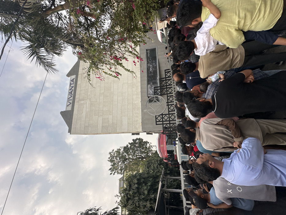 An outdoor scene in Vadodara, India, depicts a large crowd gathered in front of the Vivanta hotel. The multi-story Vivanta building, constructed of concrete with a modern facade, features its prominent "VIVANTA" signage on the rooftop. The lower section of the building exhibits dark grey paneling and a distinctive geometric metalwork design, including a circular motif.
Approximately 50 to 70 individuals, predominantly adult males, populate the foreground and midground. The crowd is densely packed, with most individuals facing and looking upwards towards the hotel building, suggesting they are observing an event or awaiting an arrival/departure. A man in a light blue shirt in the left foreground looks directly upward. Another man on the right carries a young child on his shoulders, while a different man in the center holds an infant.
To the left of the hotel, behind what appears to be a barrier or railing, a red bus partially displays "ROYAL CHALLENGERS BANGALORE" branding. A white van is partially visible further right, obscured by the crowd. Lush green trees, including a tall palm tree, and flowering bushes with pink blossoms flank the scene and are visible in the background. The sky overhead is overcast with scattered clouds.