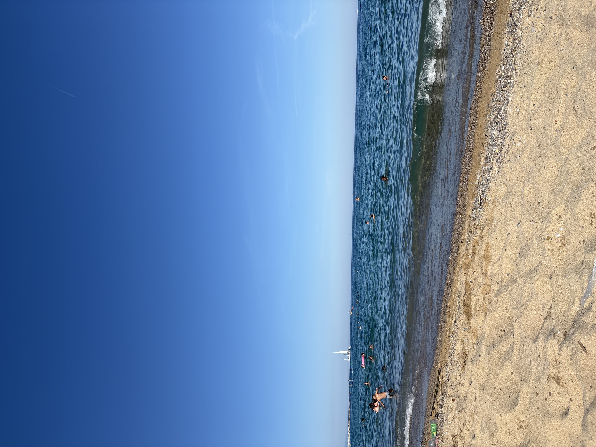 The image shows a beach scene, likely in Barcelona, Spain, based on the general appearance of the coastline and the presence of beachgoers. The view is primarily composed of the sky, sea, and beach.
The sky occupies the left portion of the image, appearing bright blue with minimal cloud cover. A thin contrail, possibly from an aircraft, is visible in the upper-left.
The sea occupies the middle section, displaying a range of blue shades indicating varying depths and water conditions. People are seen swimming and standing in the water. There is also a sailboat visible in the distance.
The beach is to the right, exhibiting a transition from pebbles near the water's edge to sand further inland. Several indentations in the sand suggesting human presence or footprints. There is a discarded green bottle.
