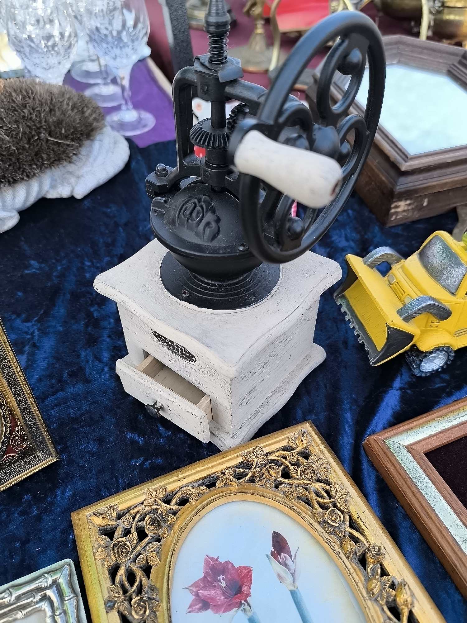 The image shows a collection of items laid out on a dark blue velvet cloth, likely at an outdoor market or flea market. The central focus is an antique-style coffee grinder with a cream-colored wooden base and a black metal top mechanism featuring a large, ornate grinding wheel with a wooden handle. In front of the coffee grinder, a small drawer is partially open.
To the left of the coffee grinder, there are three clear crystal wine glasses arranged in the background. Adjacent to the glasses is a textured, brown, hedgehog-shaped object resting on a white surface.
To the right of the coffee grinder is a small, yellow toy bulldozer with a black scoop. Further to the right and behind the bulldozer, a wooden box with an octagonal mirror is visible.
In the foreground, several ornate picture frames are arranged. The largest is a gold-colored frame with intricate floral carvings surrounding an oval picture of two red flowers. To its left and partially visible is a silver-colored frame with a bamboo-like design. To the right of the gold frame is another wooden frame with a light-colored inlay.
The overall lighting suggests it is daytime. The setting appears to be a stall at a market. While no specific landmarks of Klaipėda, Lithuania are visible, the items suggest a possible flea market or antique fair scenario within the city. There are no people depicted in the image.