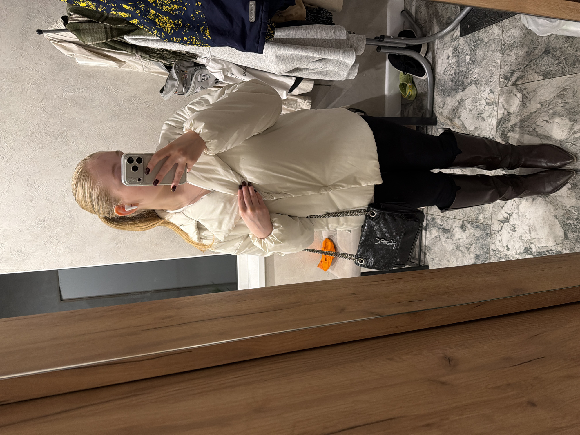 A woman is seen in a mirror selfie, wearing a white puffer jacket, black pants, and brown knee-high boots. She is holding a smartphone in her right hand, obscuring part of her face. A black handbag with a chain strap hangs off her shoulder. The mirror reflects a cluttered closet area, including clothes on a clothing rack and shoes on the floor. The reflection also reveals a grey-tiled floor. The wood frame of the mirror is visible in the foreground. Given the description of the city, it is possible this image was taken in a private residence in Rossiyskiy, Russia.