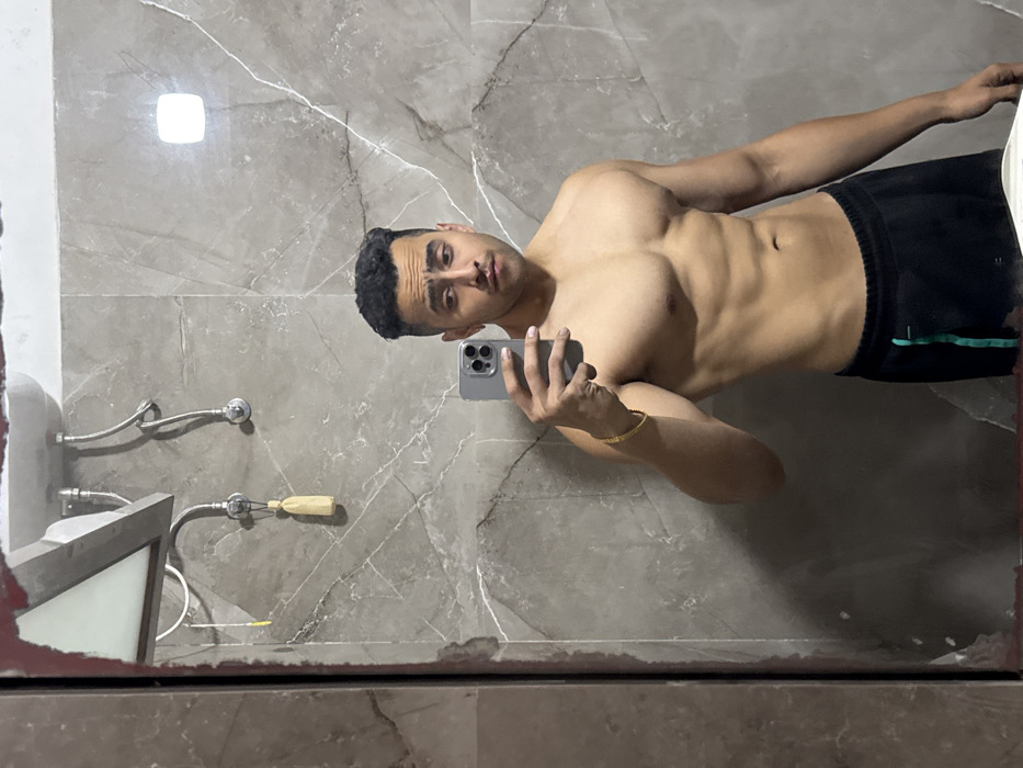 A shirtless, muscular man takes a selfie in a bathroom mirror. He has short, dark hair and is wearing black shorts with a green stripe. The bathroom has gray marble walls and a sink with exposed pipes and a cleaning brush. The mirror has visible wear and tear along the edges.
