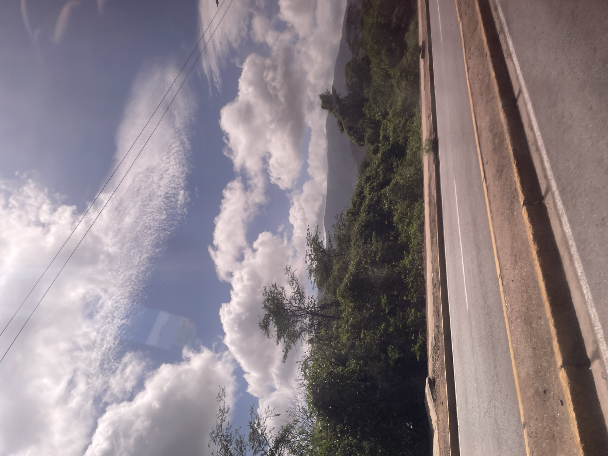 An eye-level outdoor medium shot captures a highway with a dirt shoulder and a green, overgrown embankment. The road is paved with asphalt and has a white line painted down the center, with yellow lines marking the shoulders. The embankment is covered with dense green foliage, with several trees scattered throughout. In the background, mountains rise into a sky filled with large, fluffy white clouds. Two dark lines, possibly power lines, stretch across the sky. The scene is well-lit, suggesting it was taken during the daytime.