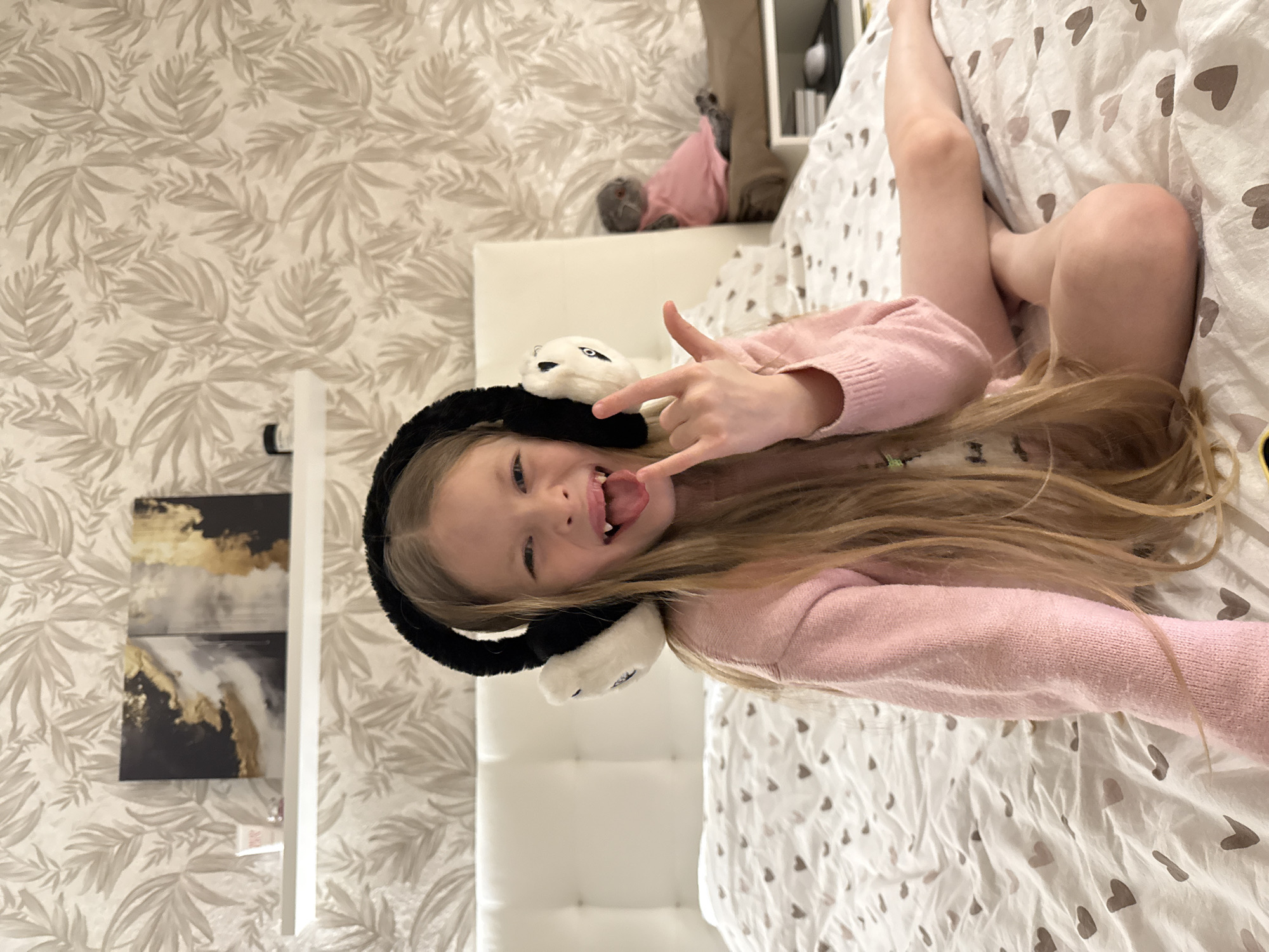 A young Caucasian girl with long blonde hair is lying on a bed with white heart-patterned sheets. She is wearing a light pink long-sleeved shirt and black and white panda earmuffs. Her legs are crossed and raised. She is making the "rock on" hand gesture with her right hand and sticking her tongue out playfully. The background includes a white headboard, wallpaper with a leaf pattern, and a white shelf with black and gold artwork. The location is a bedroom, presumed to be inside a home, but there is no indication of it being in Izhevsk, Russia.
