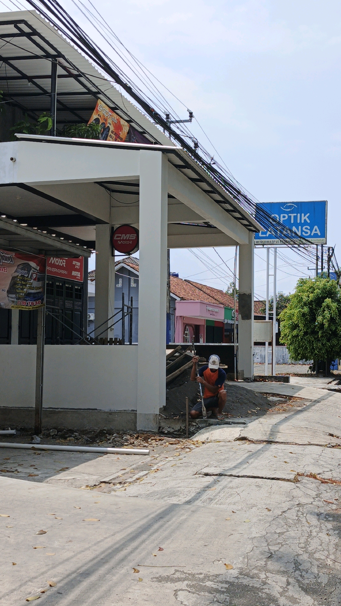 A person wearing an orange shirt, blue shorts, and a white cap is crouching on a dirt pile with a shovel in their hands. They are in front of a white pillar that is part of an outdoor structure with a corrugated metal roof. To the left of the pillar, there is a building with a sign that says "CMS Moto2" in a red circle. Further left, a banner with the word "Motog" is visible. In the background, there are several buildings with tiled roofs, including one painted pink. A blue sign with "OPTIK LATANSA" is also visible in the background. The ground is paved with concrete and dirt, with some fallen leaves scattered around. The scene appears to be in Desa Wonoharjo, Indonesia, likely an outdoor commercial area or street.