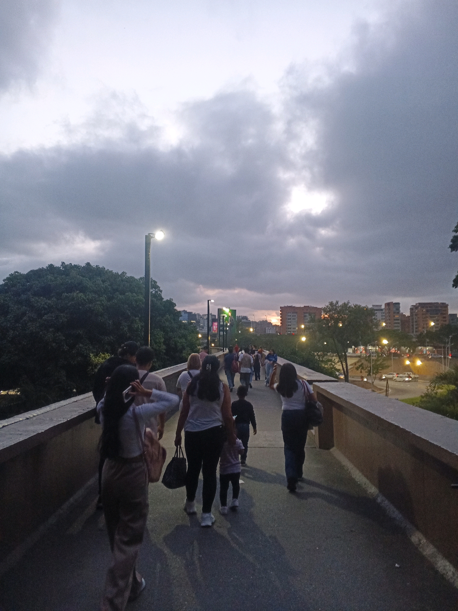 A group of people are walking on a pedestrian bridge over a road in Caracas, Venezuela. The sky is cloudy with a hint of sunlight peeking through. There are trees and buildings in the background, and streetlights are illuminated on the bridge. Some of the people are carrying bags.