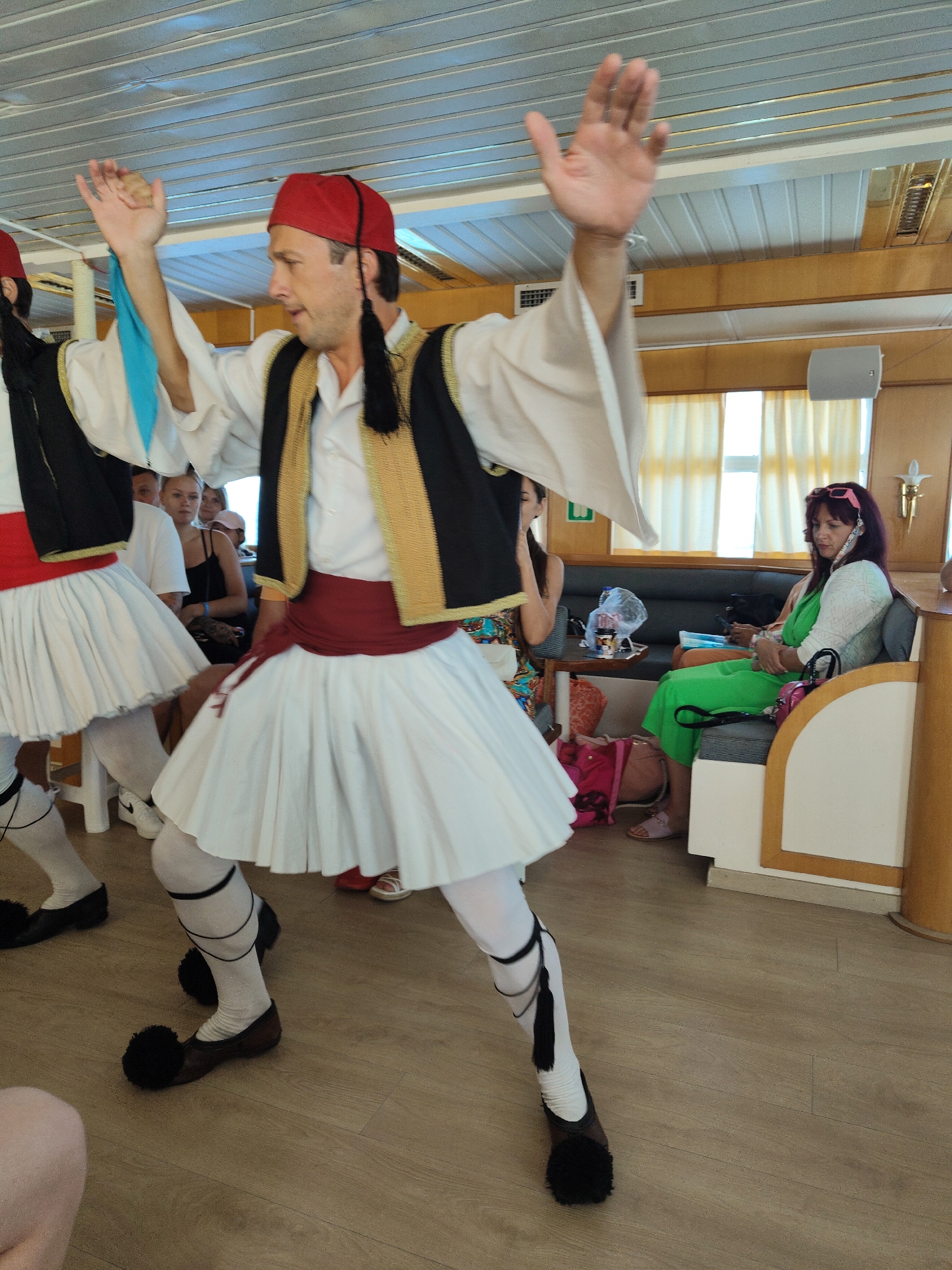 A male dancer in traditional Greek attire, a red fez, a white shirt with wide sleeves, a black vest with gold trim, and a white pleated skirt (fustanella) is performing on a wooden floor indoors. He is wearing white socks and black shoes with large black pom-poms. His arms are raised. Another dancer in similar attire is partially visible to his left. In the background, several women are seated on a bench. One woman on the right wears a green dress and pink sunglasses. The environment appears to be the interior of a boat or ferry, with a paneled ceiling and windows with yellow curtains visible. The location is described as Unknown, Greece.