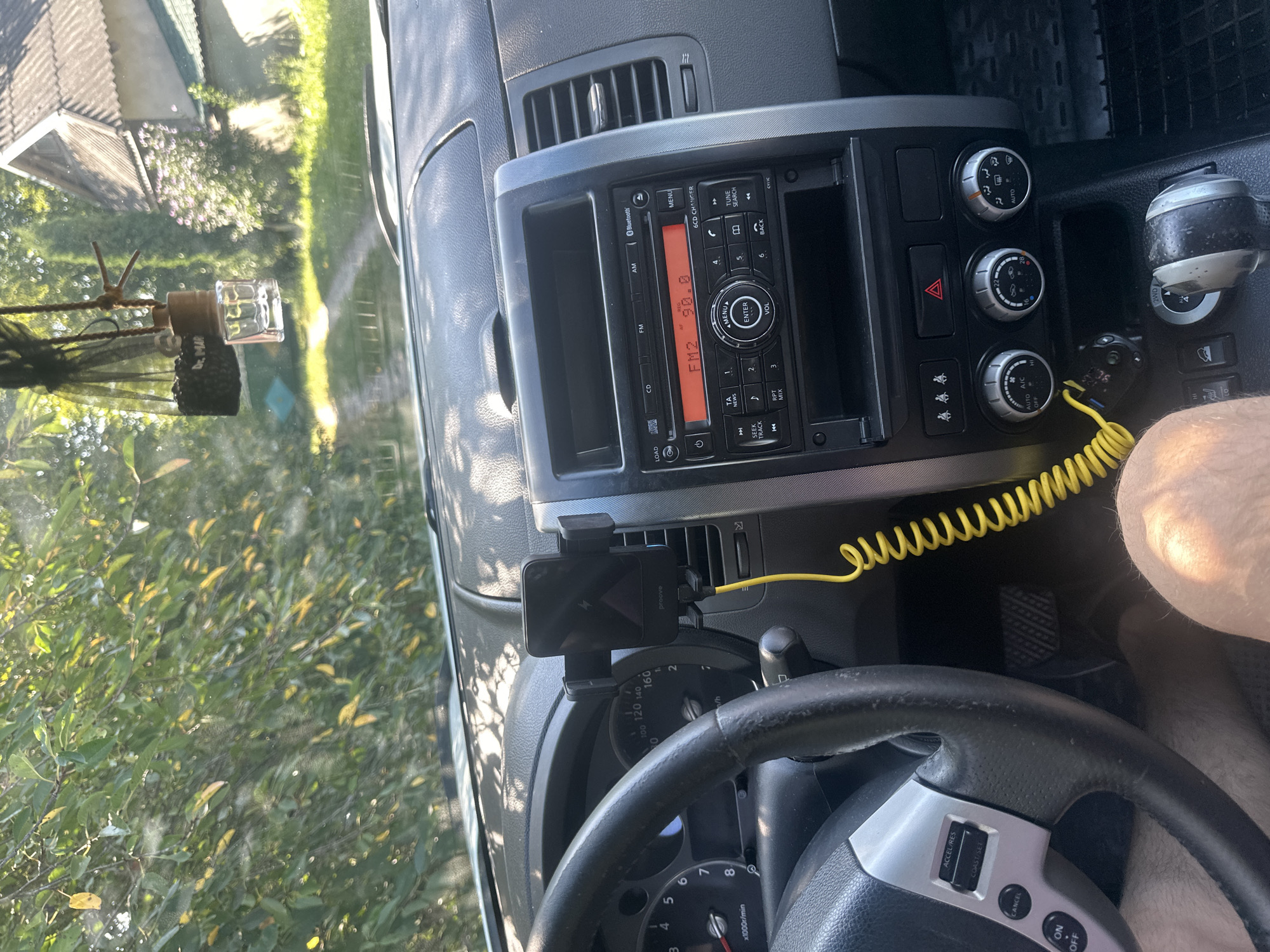 The image shows the interior of a car, likely parked on a rural road in Shatava, Ukraine, given the outdoor scenery visible through the windshield and side windows. The dashboard features a car stereo displaying "FM2 90.0", a mobile phone mounted on a holder, and various control knobs and buttons. A yellow coiled cable is connected to the phone holder and extends downwards. The steering wheel is visible on the left, with cruise control buttons. The background shows greenery, trees, and a rustic building, suggesting a rural or semi-rural setting. There is no clear indication of any people inside the car. The overall scene depicts a static moment inside a vehicle.
