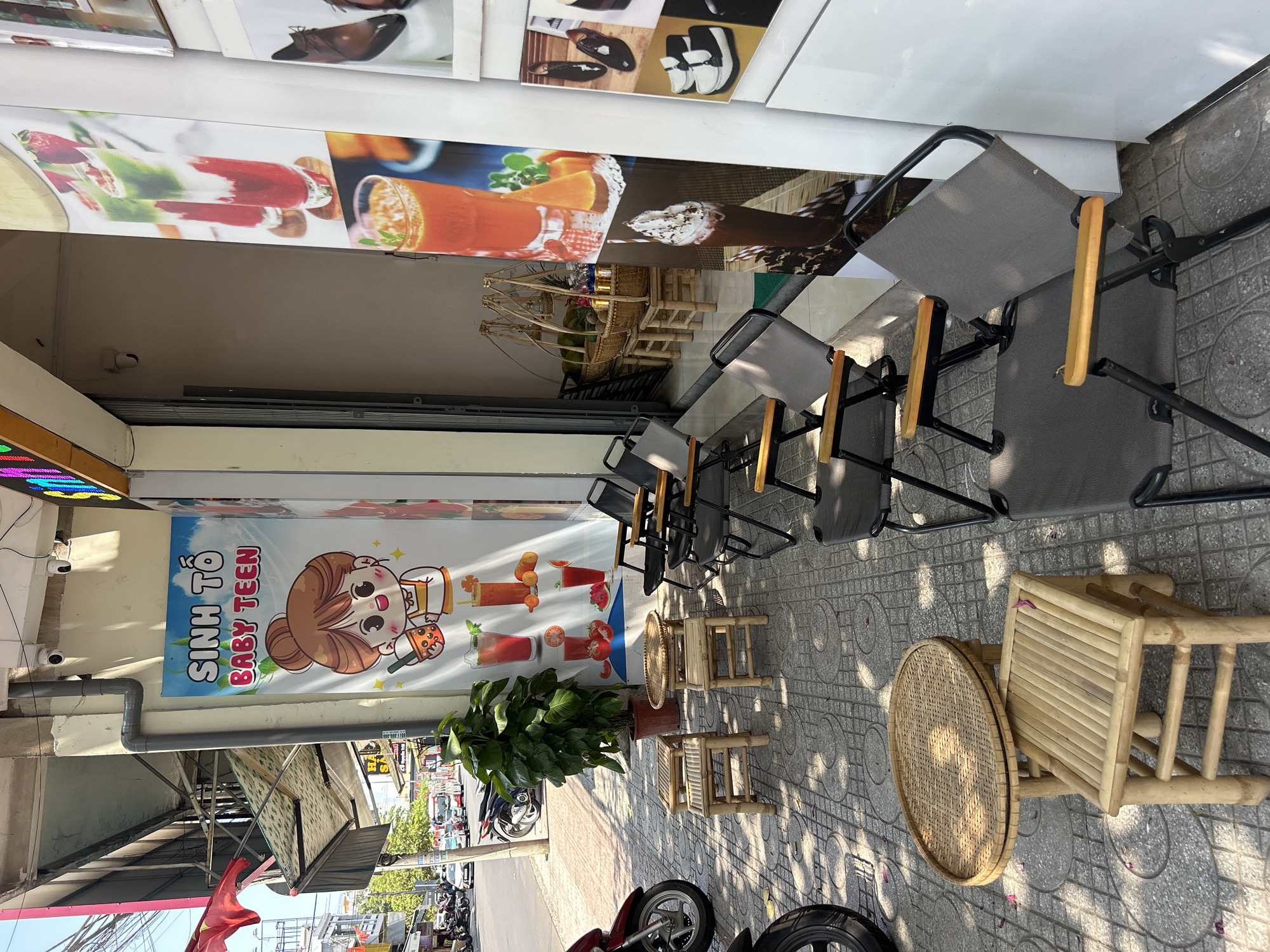 A sidewalk scene in Nha Trang, Vietnam shows an outdoor seating area for a shop. The sidewalk is paved with patterned tiles. Metal framed chairs with grey seats and wooden armrests are arranged in a row against a building. A small bamboo table and stools are nearby. A potted plant sits in front of a shop’s advertising banner, which features a cartoon girl holding a bubble tea. The banner also includes the name of the shop: "SINH TỐ BABY TEEN." Scooters are parked on the street visible beyond the sidewalk. The shop's wall has advertising posters with pictures of beverages and shoes. The weather is sunny, and the scene appears to be daytime.