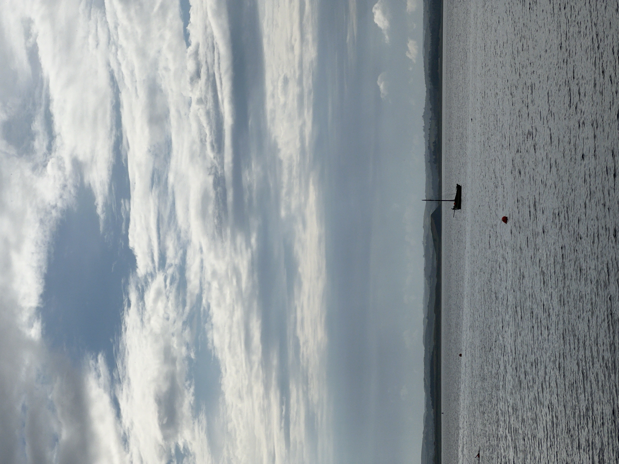 The image depicts a wide, calm body of water under a cloudy sky. In the distance, a silhouetted sailboat with a single mast is visible on the left side of the frame. The water is dark and slightly rippled, reflecting the overcast sky. The background shows a distant, hazy coastline with rolling hills. Two red buoys are present in the water, one closer to the foreground on the right and another further back near the center. The overall impression is one of a serene, possibly cool, day. The scene is consistent with views of the bay or estuary in or near Mountcharles, Ireland, a coastal town.