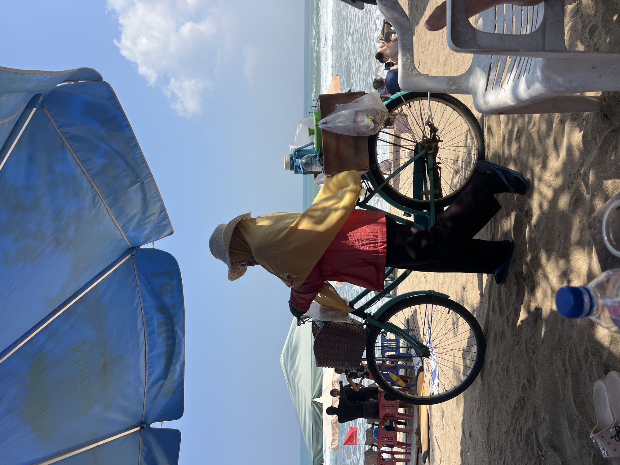 In Kuta, Indonesia, on a beach under a blue umbrella, a vendor pushes a teal bicycle equipped with a wooden box on the back. The box contains a blue thermos, a light green container, and items in plastic bags. The vendor, wearing a straw hat, yellow scarf, and red jacket, has their back to the camera. They are walking on the sand towards the ocean, where waves are breaking and a few people are visible in the water. Beach chairs and umbrellas are scattered on the sand.