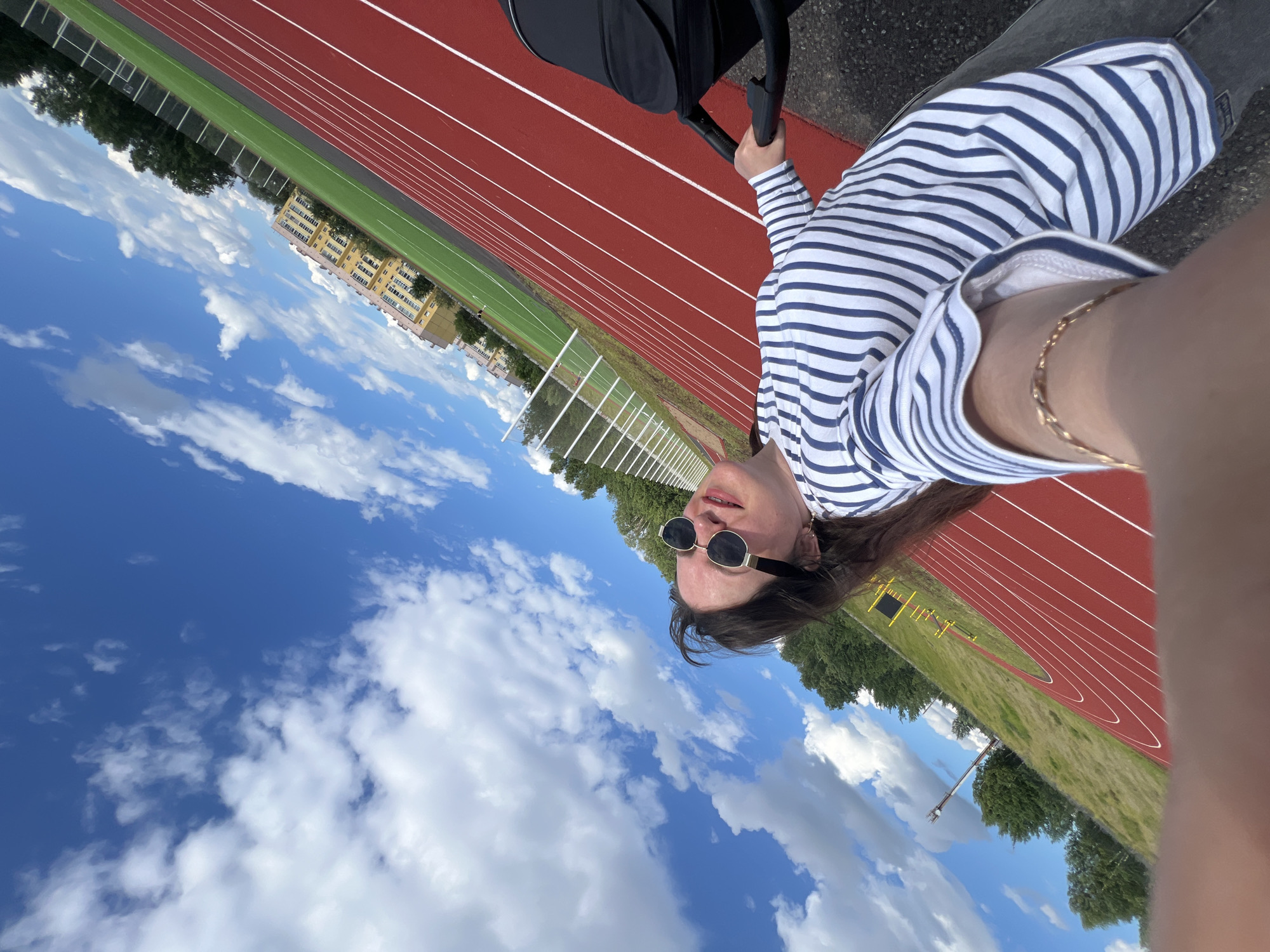 In this photograph, a person wearing a blue and white striped long-sleeved shirt and black sunglasses is captured in a selfie. They are holding onto the handle of what appears to be a stroller, suggesting they might be pushing it or have just stopped. The setting is a red running track with white lane markings, bordered by a green field. Beyond the field, there are trees and a multi-story yellow building. The sky is a vibrant blue with scattered white clouds, indicating a clear and sunny day. The image is taken at an angle, with the person's face and the stroller in the foreground and the track and surrounding environment extending into the background. The person's expression is neutral. The time of day appears to be daytime, likely late morning or afternoon, given the brightness of the sun. The location is identified as Noviy, Russia.