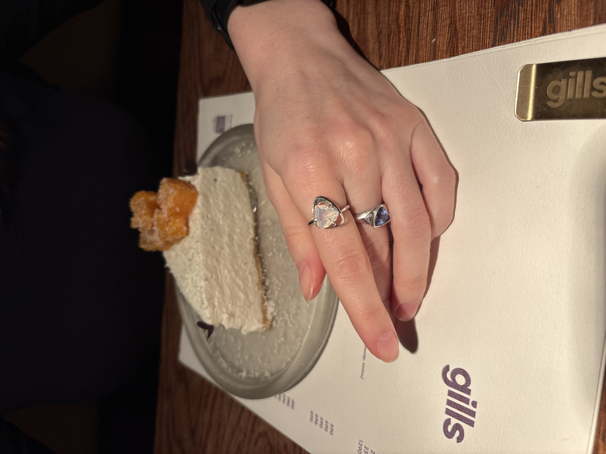 Here is a detailed description of the image:
**Scene:** The image depicts a table setting in a restaurant. Dominating the scene is a person's hand adorned with two rings, positioned next to a plate with a slice of cake and a menu card. The environment suggests an indoor dining experience.
**Objects:**
* **Hand:** The hand is light-skinned and has two rings on the ring and middle fingers.
* **Rings:** One ring features a large, white stone, while the other has a smaller, blue stone.
* **Cake:** A slice of layered cake is on a clear plate with a garnish on top.
* **Menu:** A white menu card with the restaurant name "gills" printed in purple is visible. A gold-colored metal bar also with the restaurant name is located in the upper-right corner of the menu.
* **Table:** The table has a wooden surface.
**Text on Menu:** The menu has the text "gills" prominently displayed. Other text that appears to be prices is also visible.
**Environment:** The scene is indoors, likely within a restaurant. The lighting seems warm.
**Implied Location (Based on the menu name):** Gills restaurants is a restaurant in the city of Saint Petersburg, Russia.