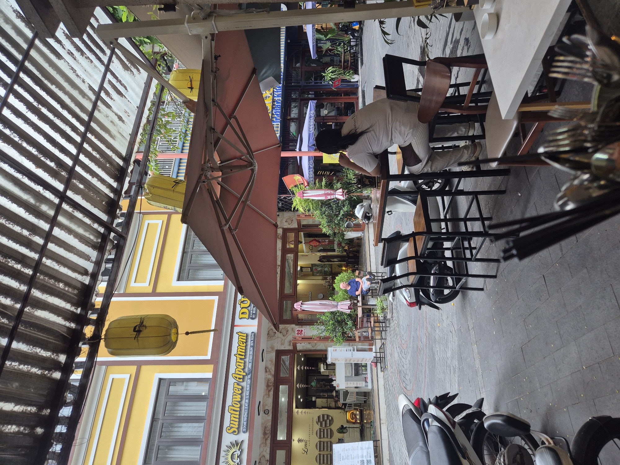The image shows an outdoor seating area of a cafe or restaurant on a street in Da Nang, Vietnam. The area is covered by a large, reddish-brown umbrella and has several tables and chairs. Two motorcycles are parked nearby. In the background, there are buildings with yellow facades and balconies, suggesting a commercial or residential street. One building displays a sign that reads "Sunflower Apartment." A man wearing a blue shirt and shorts is sitting at one of the tables. Another person, wearing a white shirt and striped pants, is seated at a different table facing away from the camera. The scene appears to be during the daytime, with natural light filtering through the overhead covering.