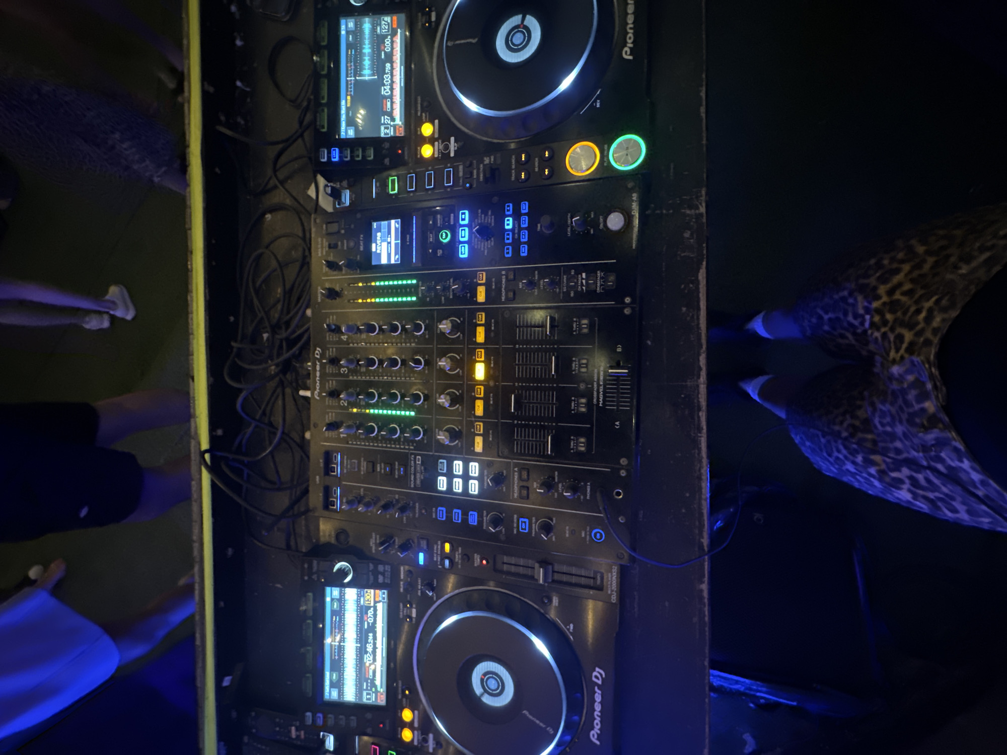 The image shows a DJ setup, likely at a club or event, with two Pioneer CDJ-3000 multiplayers and a Pioneer DJM-A9 mixer. The lighting is dim and colorful, with blue and green hues dominating. In the background, a few people can be seen standing, their legs visible. One person is wearing a leopard-print skirt. The DJ equipment is the central focus, with its illuminated displays and controls. The scene suggests a lively atmosphere, typical of a nightlife setting. No specific location in Moscow, Russia, can be identified from the image alone.