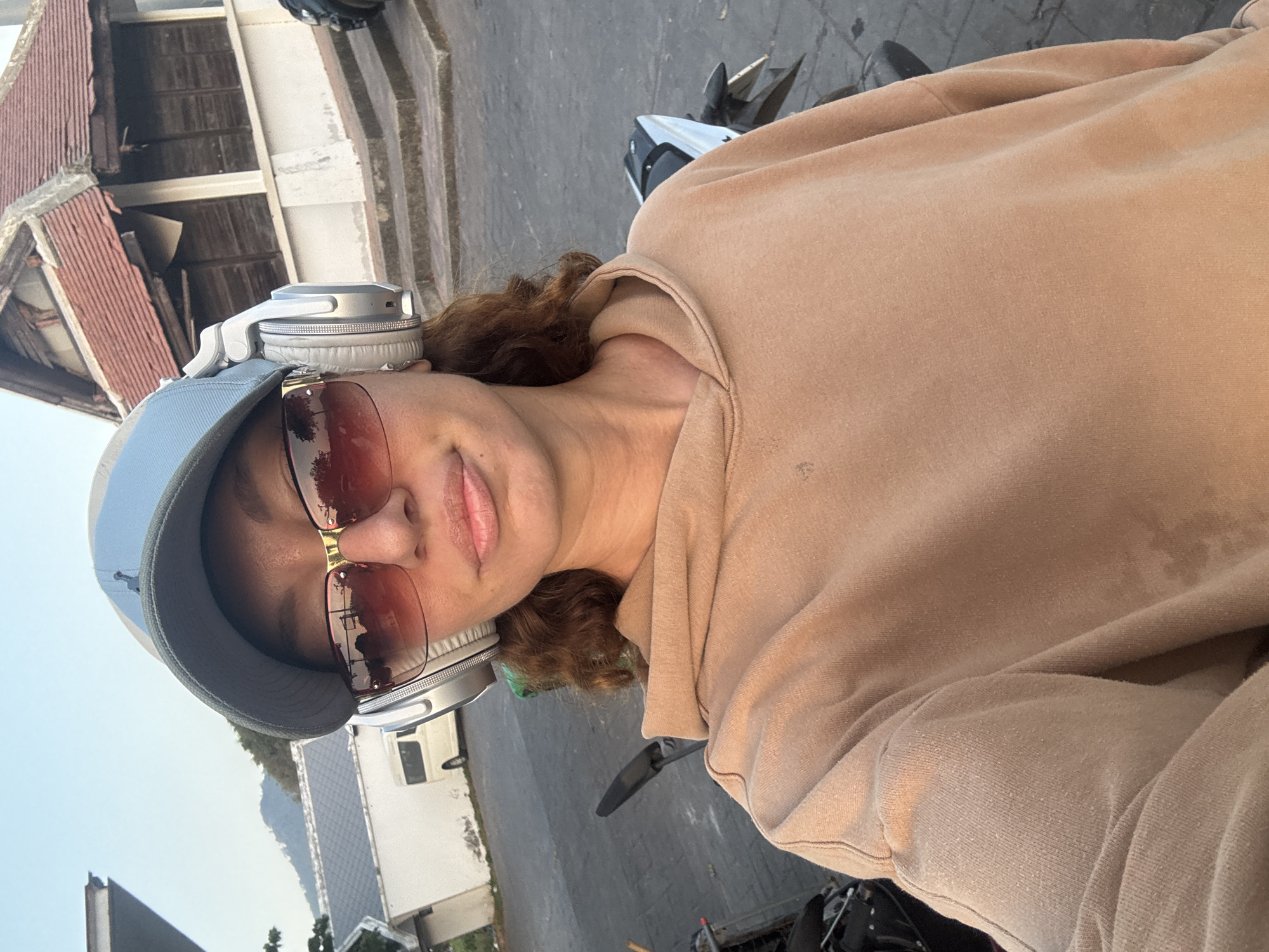 A woman wearing a light blue cap, sunglasses, and headphones is sitting on a motorcycle. She is wearing a light brown hoodie. The background shows buildings and a road in Baan Chaweng, Thailand.