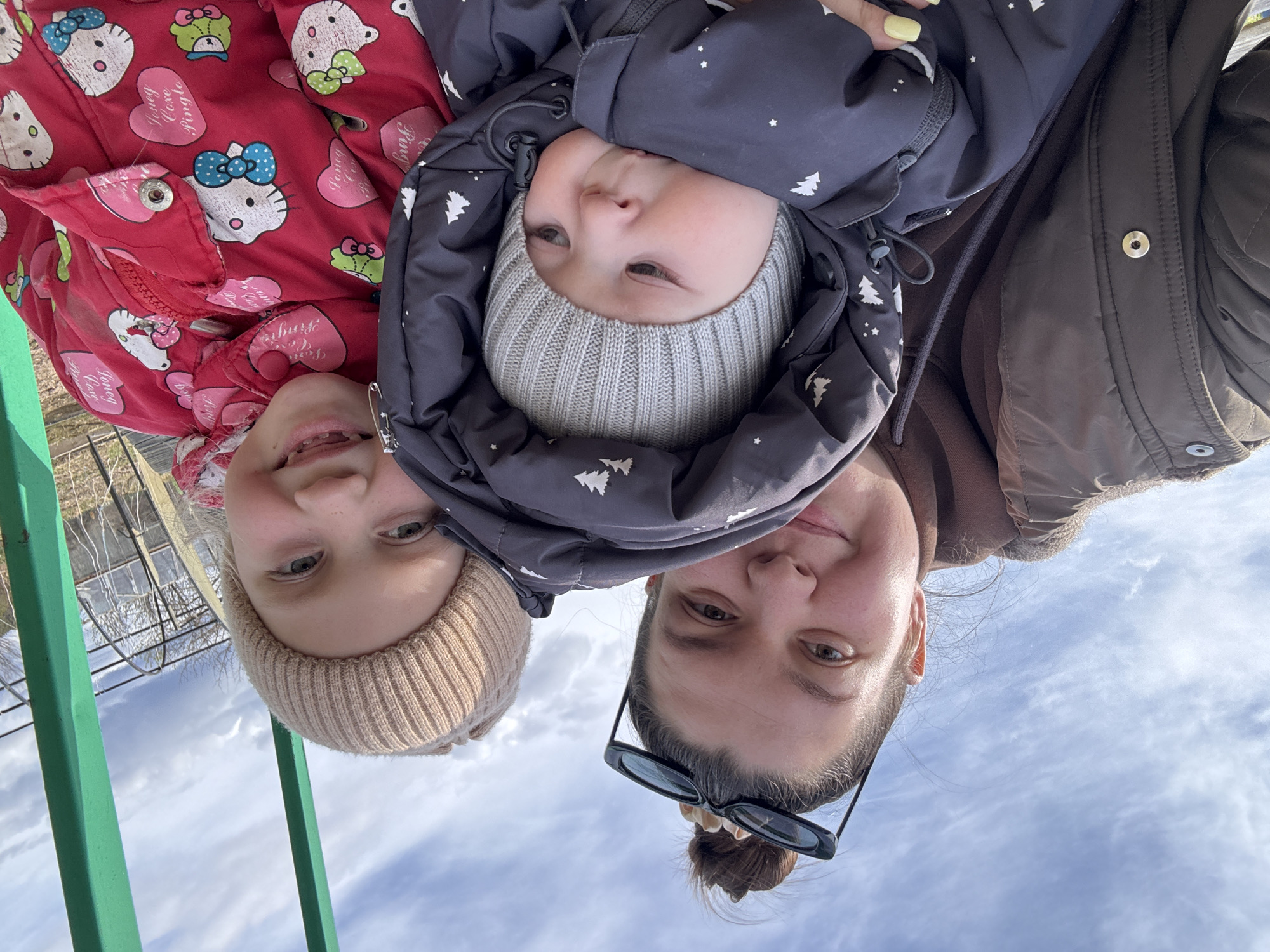 This upside-down image, captured from a low angle, features three individuals against a backdrop of a cloudy sky. On the right, an adult woman with dark hair in a bun wears sunglasses on her head and a brown jacket over a dark brown top. Her visible fingernails are painted a pale yellow. To her left, a young girl with light hair, wearing a beige knit beanie, smiles subtly, revealing braces. She is dressed in a red jacket adorned with white Hello Kitty characters and pink hearts, some of which read "Lovely Love Pringle." In the center, a baby or toddler looks curiously towards the camera, bundled in a dark gray puffer jacket with white Christmas tree patterns and wearing a light gray knit beanie.
The setting is outdoors during the day, under a sky with scattered white clouds. A bright green metal railing is visible in the foreground on the left, with hints of a wire fence and bare trees in the distance. The subjects appear calm and content, suggesting a peaceful family moment. The overall scene takes place in Lukintsy, Russia.