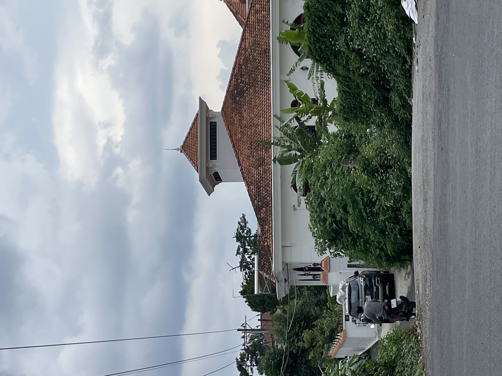 In Phường Xuân Hương - Đà Lạt, Vietnam, on an overcast day, a paved road leads to a white building with a red tile roof. A dark-colored SUV is parked in the driveway of the building. A person is riding a motorcycle away from the building and towards the viewer, on the right side of the road. The building is surrounded by lush green trees and foliage. Utility lines are visible running across the sky.