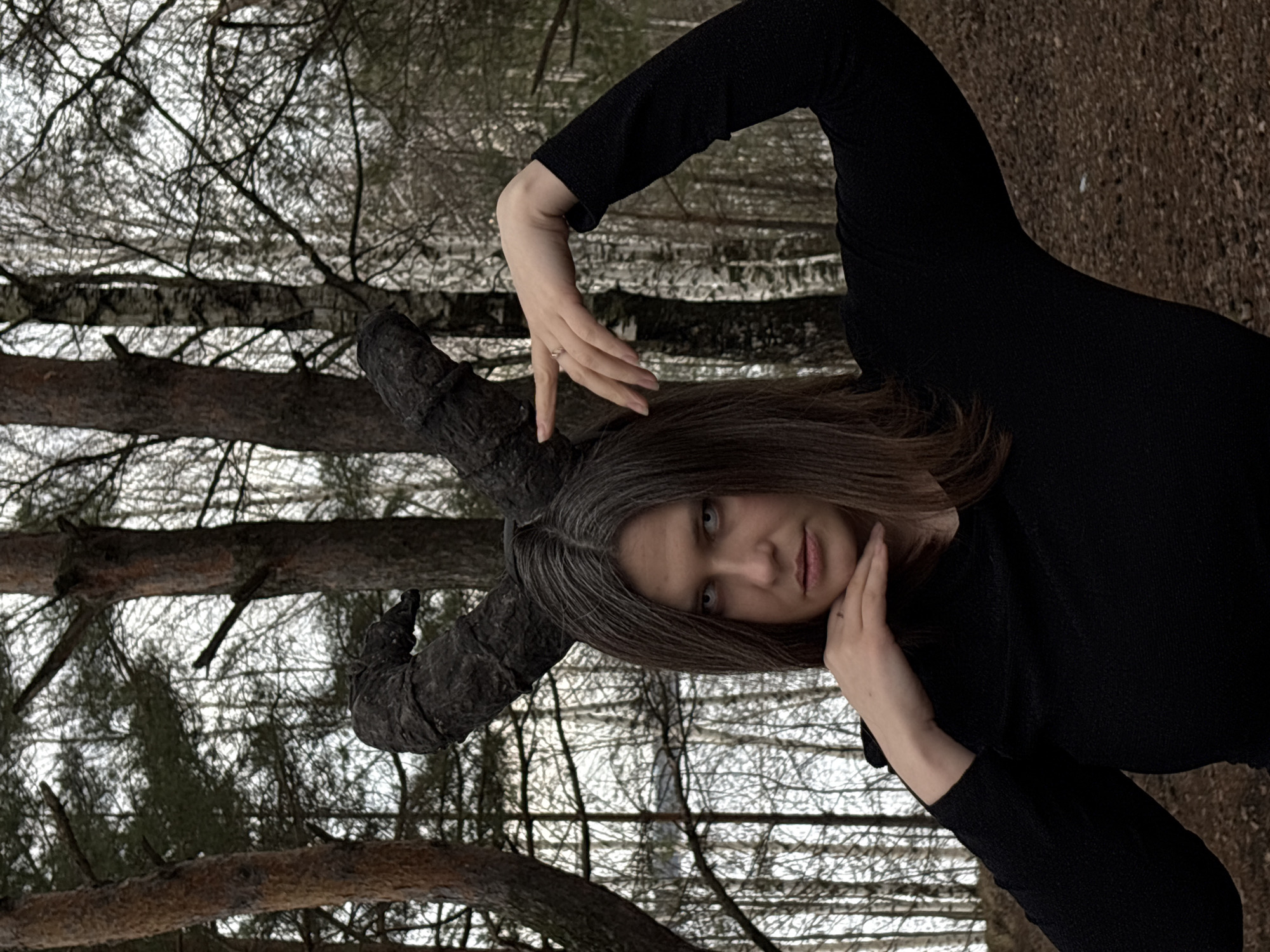 A photograph depicts a woman outdoors in what appears to be a wooded area near Chaikovsky, Russia. She has medium-length brown hair with gray roots, pale skin, and is wearing a long-sleeved black dress. She is positioned at an angle, with the wooded environment visible in the background. Above the woman is a tree with thick, dark branches resembling horns. The woman is posing with her hands near her face, in a contemplative expression. The background features a mixture of pine trees and bare trees. The ground appears to be covered in dirt or pine needles.