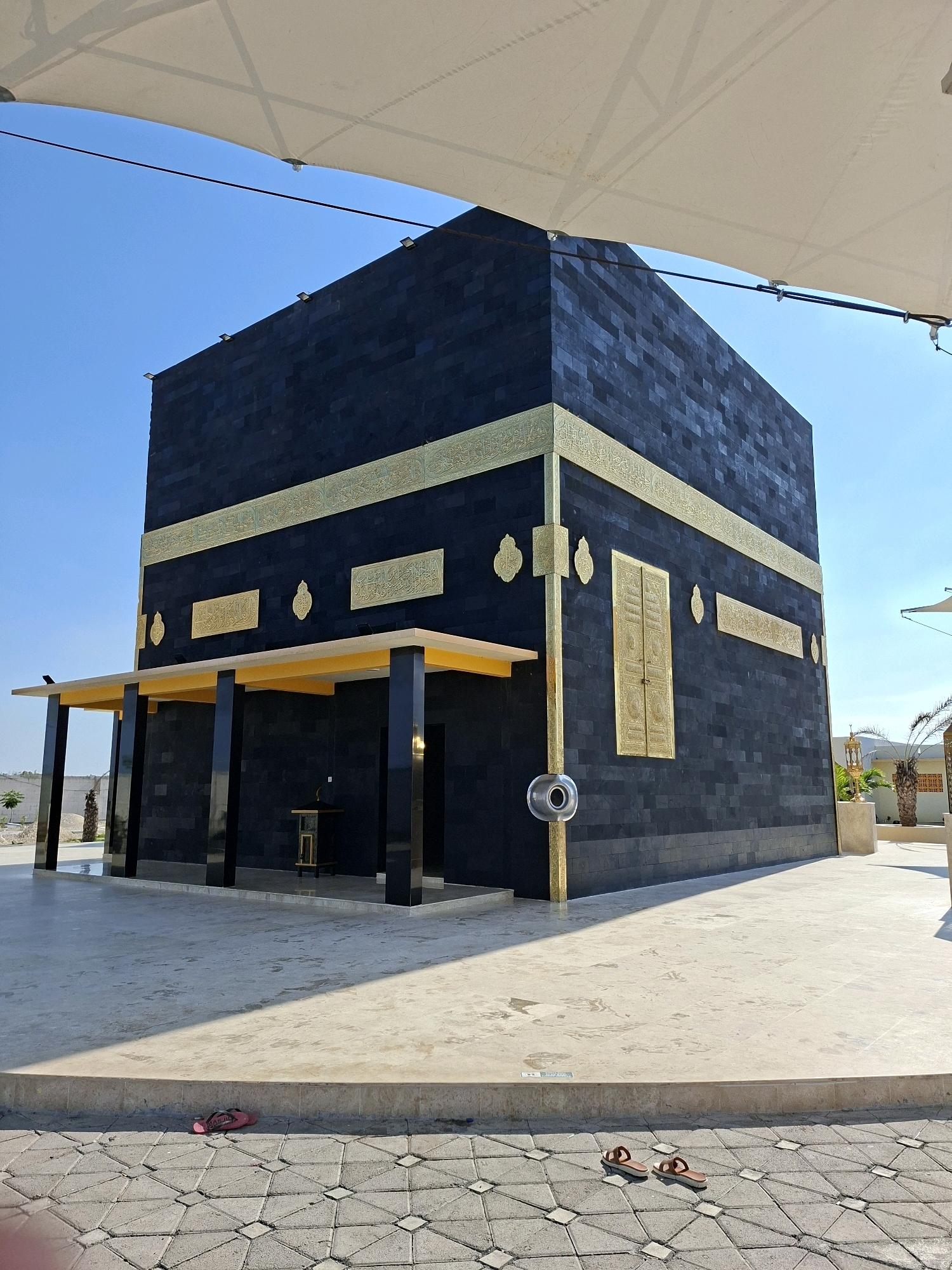 The image displays a replica of the Kaaba, an Islamic holy site, situated outdoors under a clear blue sky. The structure is predominantly black, with ornate gold embellishments along its edges and decorative panels on its sides. A golden door-like feature is visible on the right side. The Kaaba replica is surrounded by a paved area, and a light-colored canopy extends over a portion of the scene. In the foreground, two pairs of sandals are present on the paved ground. Palm trees and some sparse vegetation are visible in the background, suggesting a warm climate. The specific location is identified as Jrengik, Indonesia.