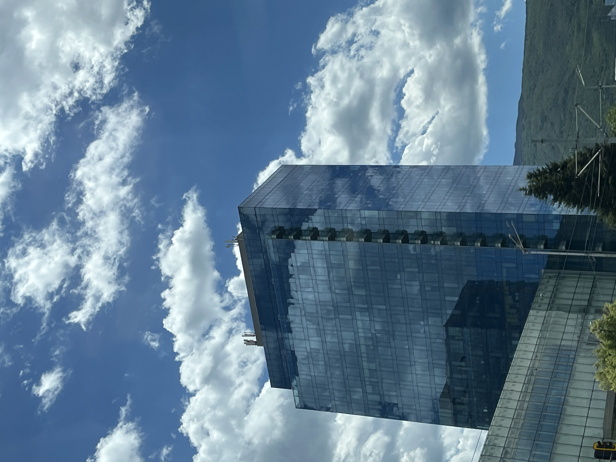 An exterior, medium shot captures a modern, glass-faceted skyscraper in an urban environment under a bright, partly cloudy sky. The building, predominantly blue-tinted glass, reflects the surrounding environment, including the sky and clouds. The skyscraper is tall and rectangular, with visible horizontal divisions between floors and vertical elements on one side. In front of and to the left of the skyscraper is a lower, also glass-covered building, with its own set of windows. At the base of the skyscraper, a dark green evergreen tree is visible. In the background, a lush, green, tree-covered mountain range recedes into the distance. The sky is a vibrant blue, dotted with fluffy white cumulus clouds. The overall impression is one of a contemporary cityscape bathed in natural light. There are no people visible in the image.