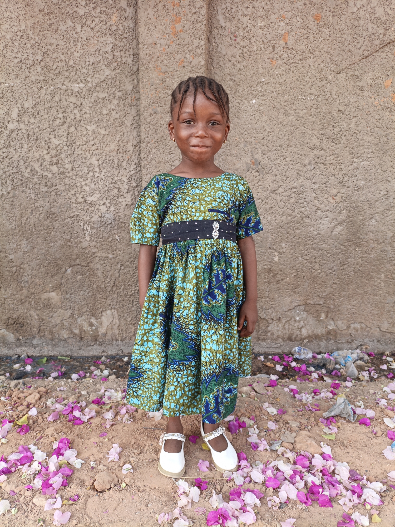 A full shot of a young girl, likely about 5 years old, stands in front of a rough, textured wall. She has dark skin, braids, and a slight smile. She is wearing a colorful dress with a green and blue pattern and a black belt with small, reflective embellishments. Her white shoes have straps across the top of her feet. The ground around her is dirt covered with pink and white flower petals, and scattered trash. The location is in Gwagwarwa, Nigeria.